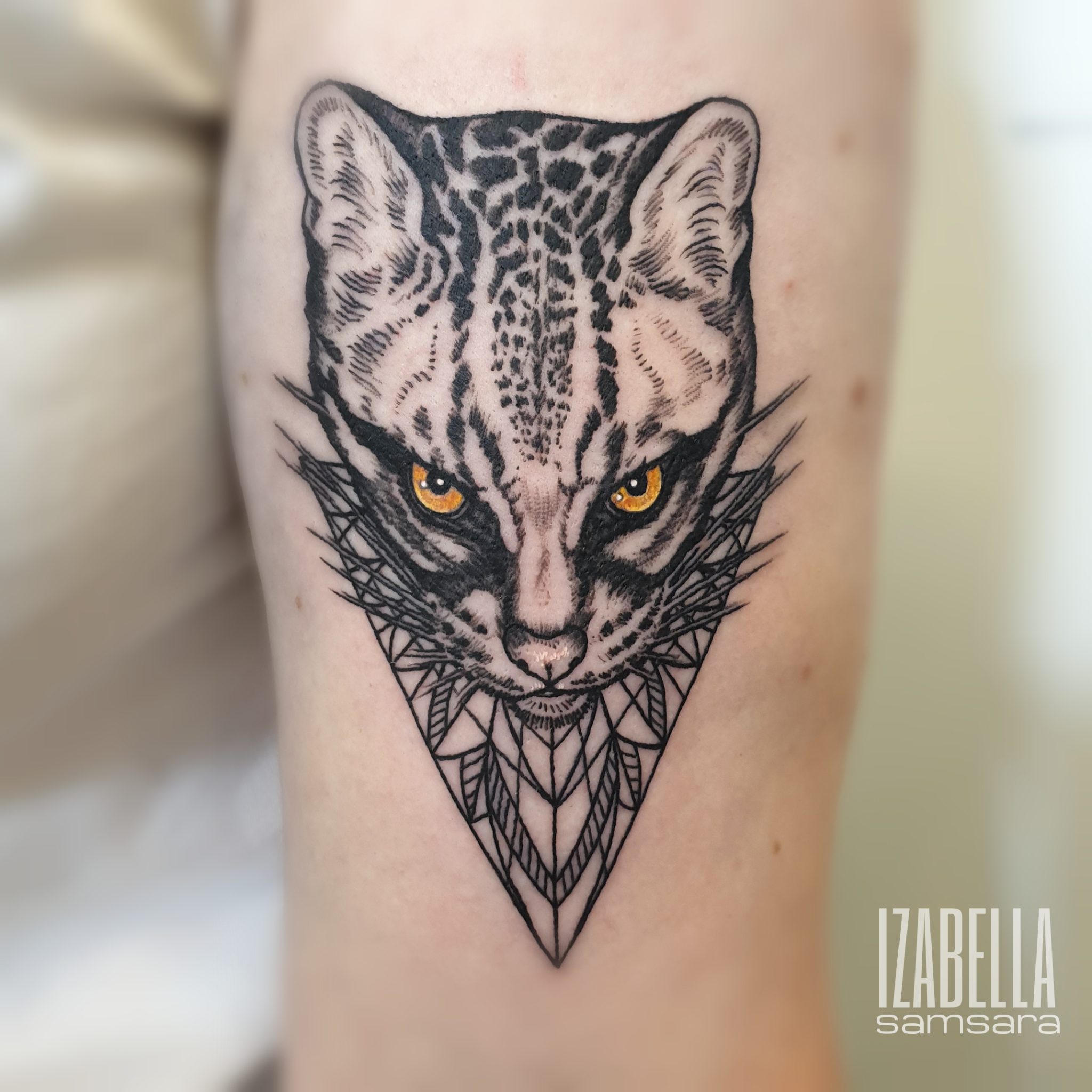 A detailed black and white tattoo of a leopard's face, featuring striking amber eyes. The design incorporates geometric shapes, creating a triangular frame around the leopard, adding depth and a modern aesthetic. The intricate linework emphasizes the texture of the leopard's fur, showcasing an artistic blend of realism and abstract patterns.