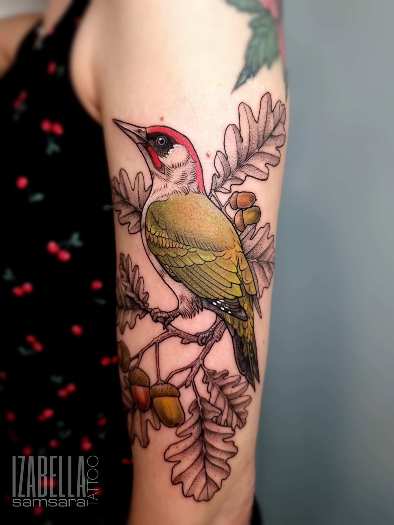 A vibrant tattoo depicting a green woodpecker perched on a branch surrounded by oak leaves and acorns. The bird features a distinctive red crown and black markings on its face, with detailed shading that highlights its feathers. The composition is rich in color, emphasizing the natural elements of the design.
