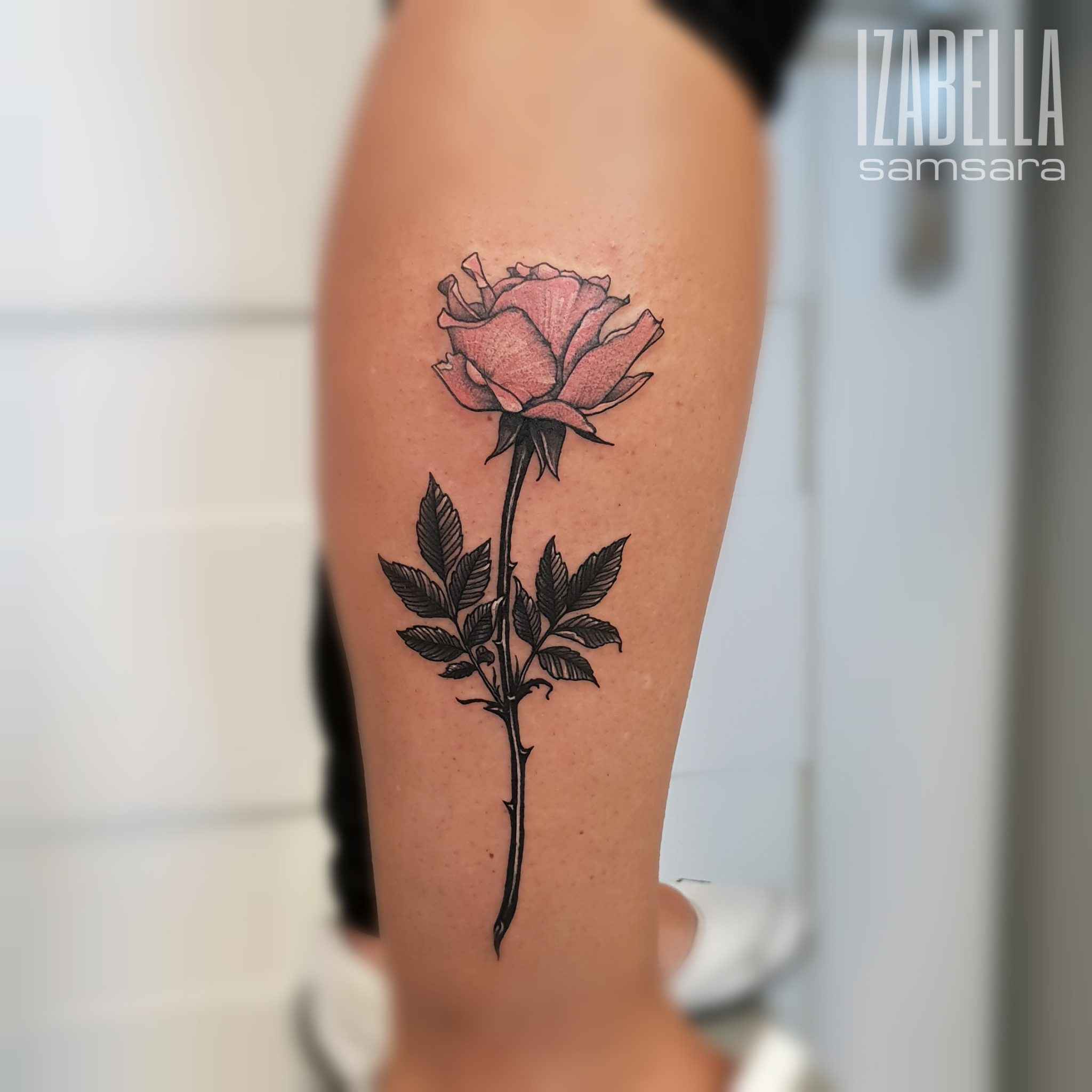 A black and gray tattoo of a rose with detailed petals and leaves, showcasing a blend of realistic and artistic elements. The rose is prominently displayed on the forearm, emphasizing its intricate design and the contrast between the dark shading and the soft tones of the flower.