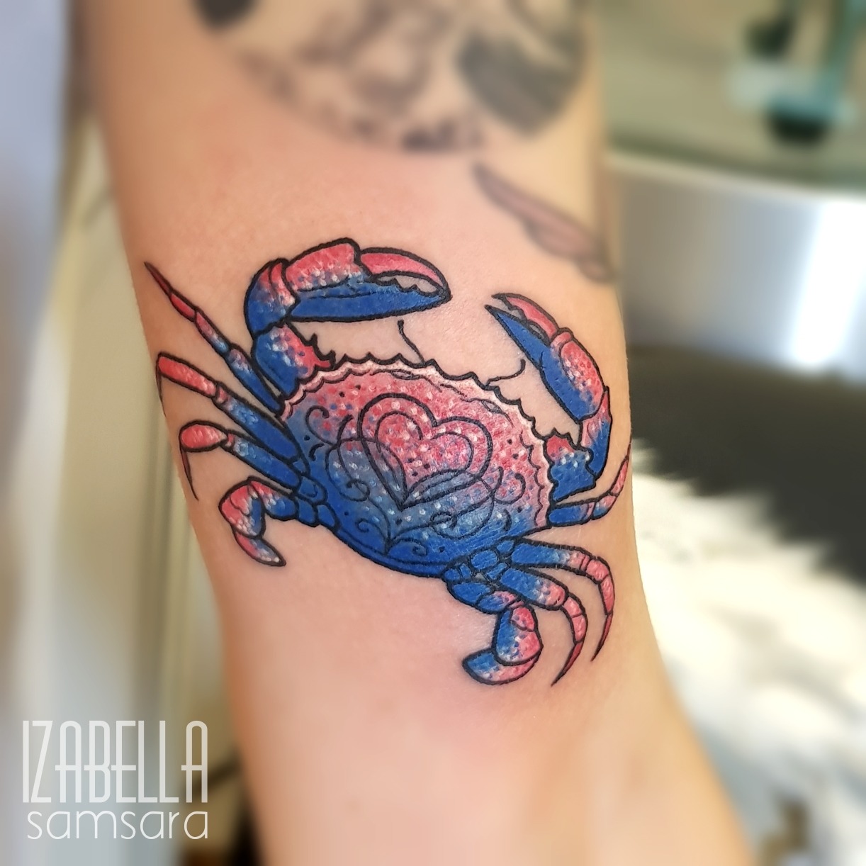 A vibrant tattoo of a crab with red and blue colors. The crab is intricately designed with decorative patterns, featuring a heart shape on its shell. The tattoo is located on the arm and showcases a playful and artistic representation of the crustacean.