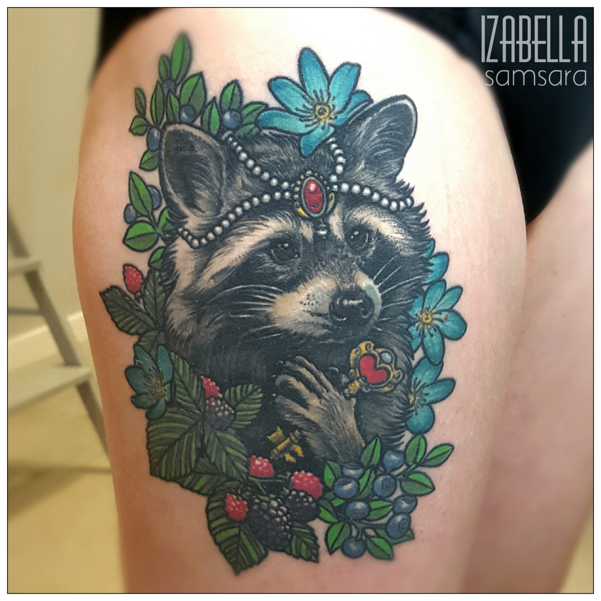 A colorful tattoo featuring a raccoon adorned with a pearl necklace and a red gemstone crown. It is surrounded by various green leaves, flowers, and berries, showcasing a whimsical and detailed design.
