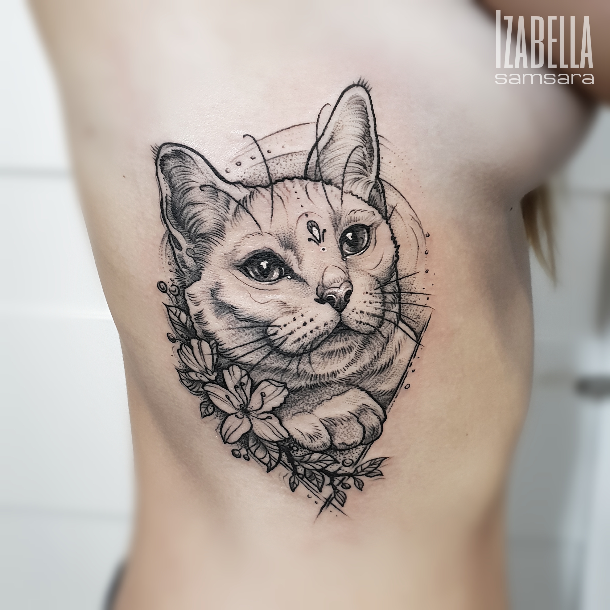 A detailed black and white tattoo of a cat's face, featuring expressive eyes and prominent whiskers. The cat is adorned with a decorative dot above its forehead and surrounded by floral elements, adding an artistic touch. The design emphasizes the cat's fur texture and facial features, making it a striking piece on the skin.