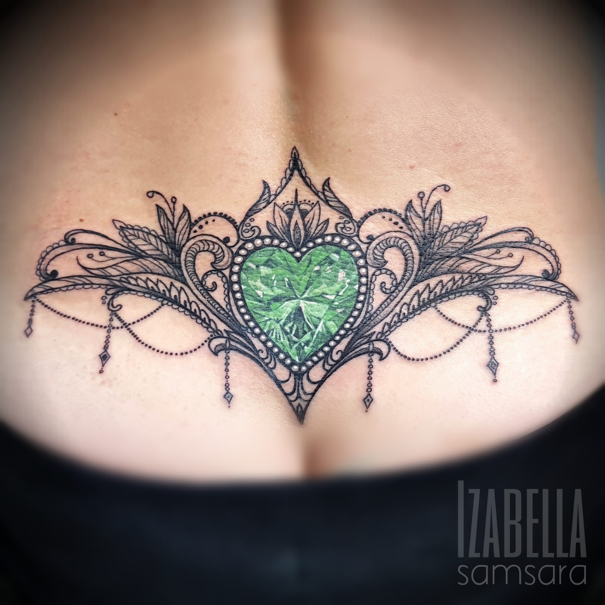 Intricate black linework tattoo featuring a green heart-shaped gem surrounded by ornate floral and decorative elements. The design includes delicate bead chains and intricate detailing, creating a stunning visual effect on the upper back.
