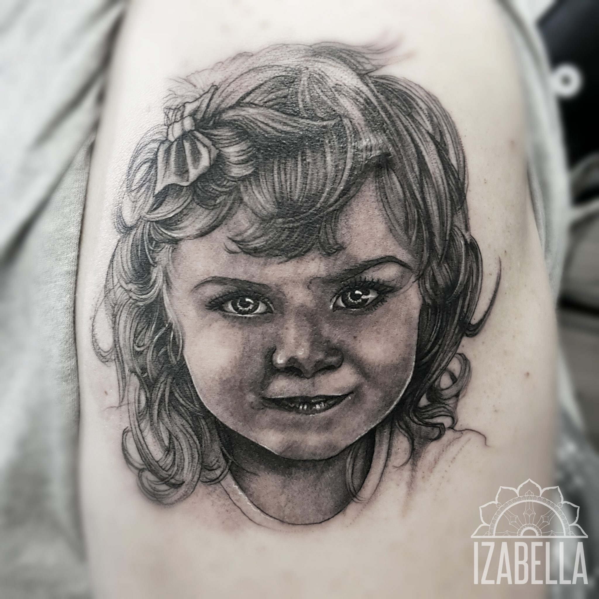 A detailed black and grey tattoo depicting the portrait of a young girl with curly hair and a bow. The artwork captures her expressive eyes and a playful smile, showcasing intricate shading and fine lines to create a lifelike appearance.