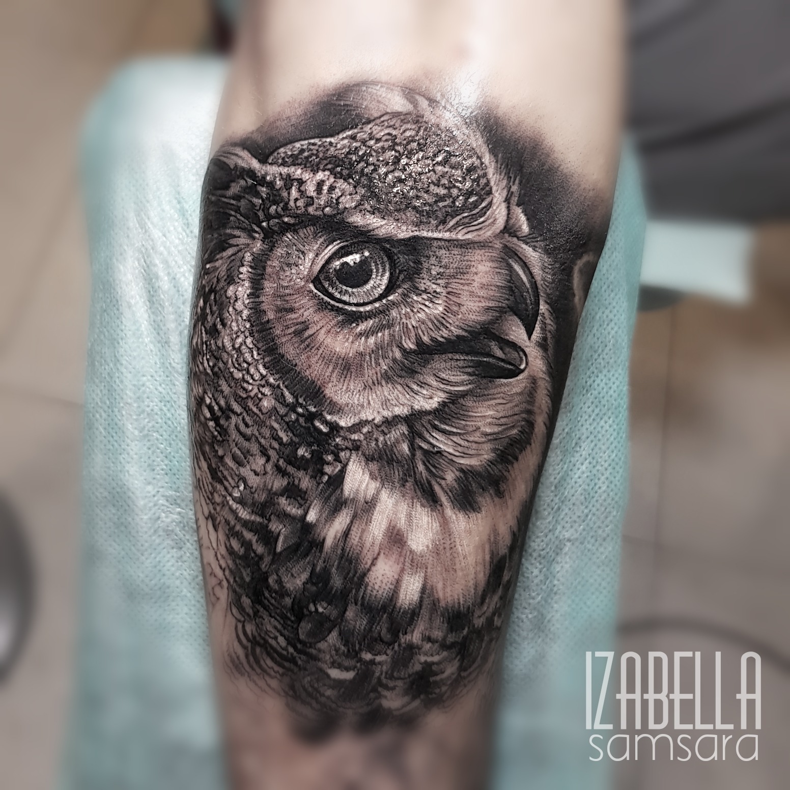 Detailed black and grey tattoo of an owl's head, showcasing intricate line work and shading to highlight the textures of its feathers and expressive eyes, positioned on the lower leg.