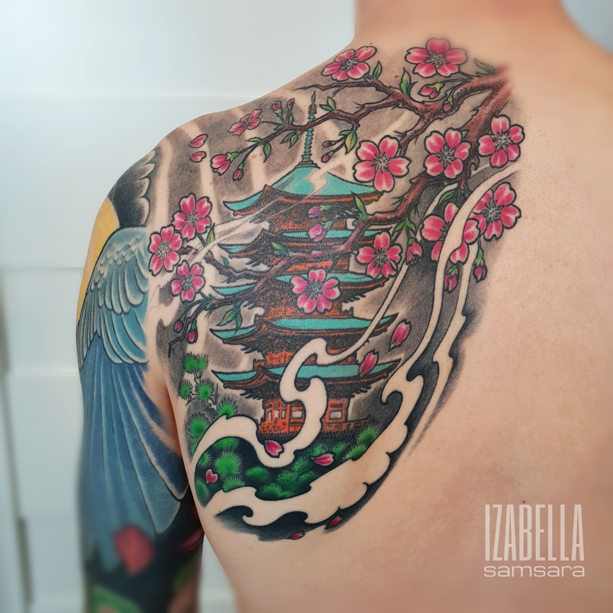 A colorful tattoo featuring a traditional Japanese pagoda surrounded by cherry blossom branches. Delicate pink flowers bloom alongside stylized clouds and lush greenery, creating a vibrant and intricate design that beautifully captures Japanese culture.