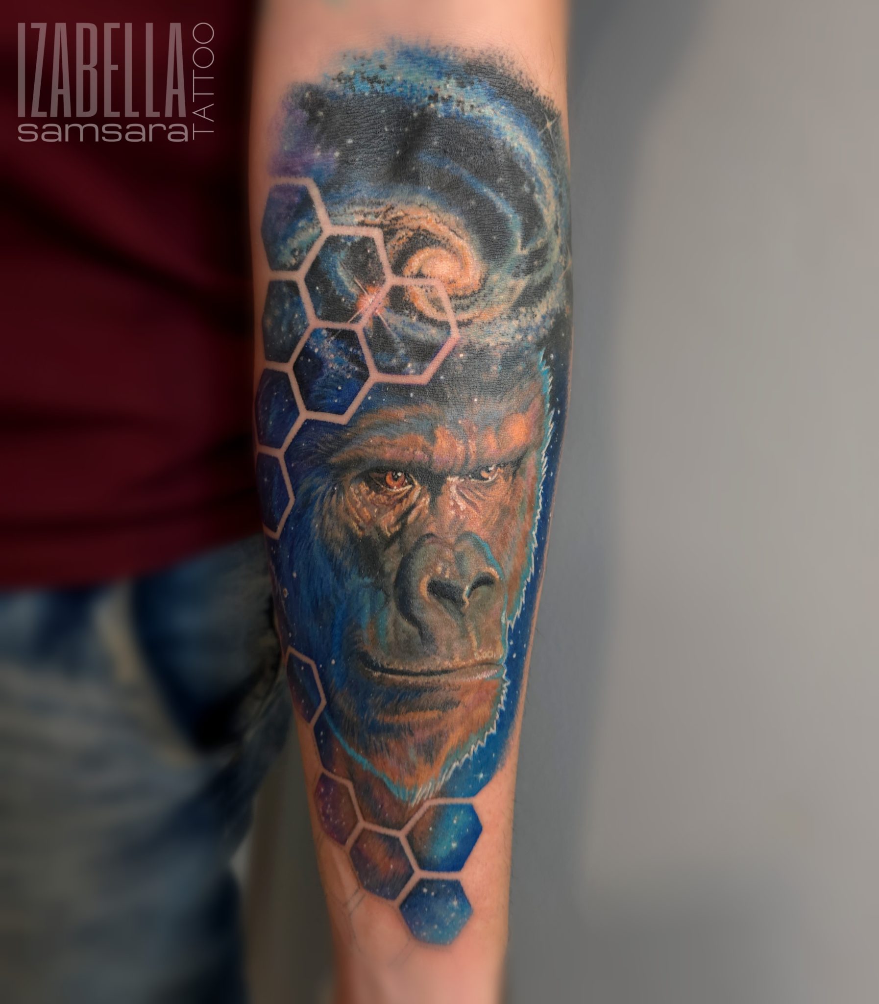 A vibrant tattoo featuring a close-up of a gorilla's face, set against a cosmic background with swirling clouds and stars. The design is accentuated with hexagonal shapes, creating a striking and modern look. The gorilla has detailed fur and expressive eyes, capturing a sense of depth and realism.