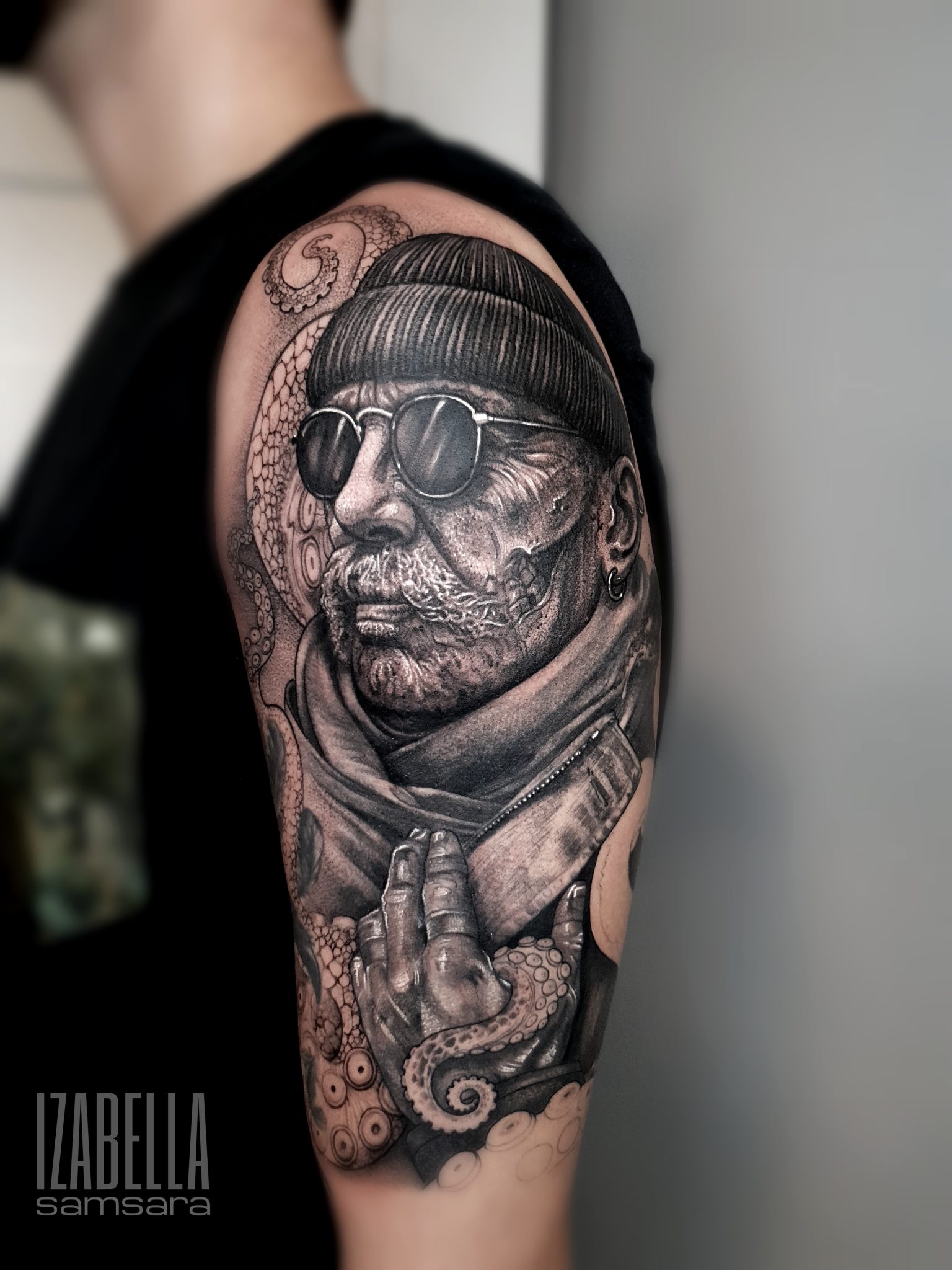 A detailed black and gray tattoo featuring a rugged man wearing sunglasses and a beanie, with a thick mustache and textured skin. He is positioned in a thoughtful pose, with one hand partially concealed in a scarf while the other hand displays a unique hand gesture. Surrounding him are intricate elements, including an octopus with visible tentacles and large, expressive eyes, adding depth and interest to the design.
