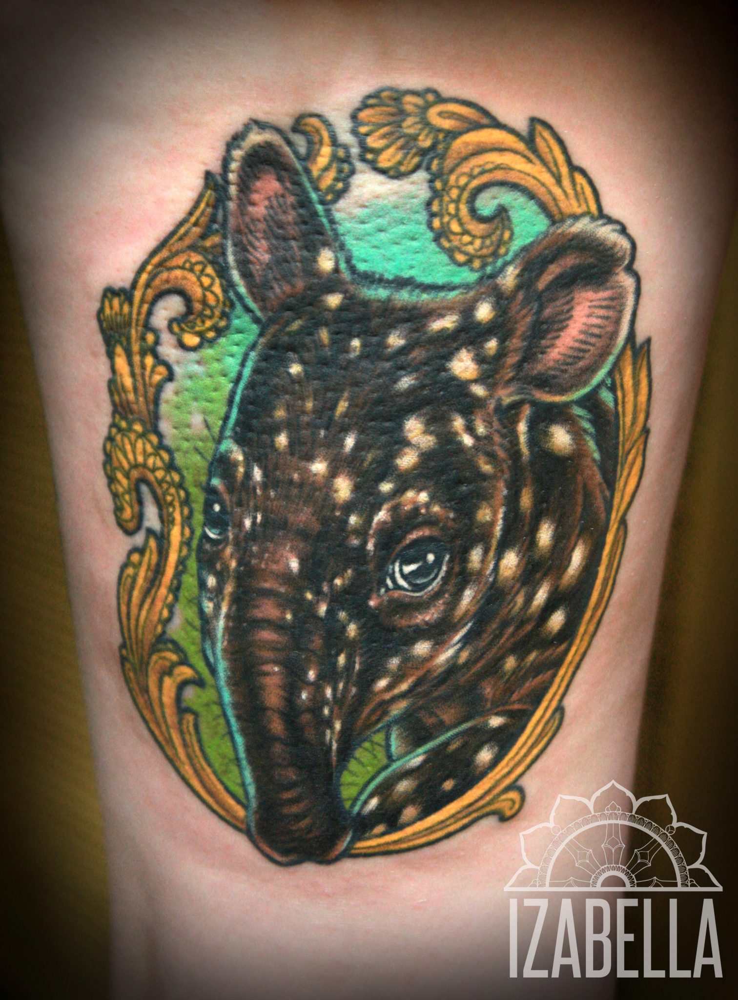 A vibrant tattoo of a young tapir's head, featuring intricate details such as its distinctive spotted fur and expressive eyes. The tapir is surrounded by an ornate golden frame with decorative swirls, set against a lush green background, creating an appealing contrast. Ideal for animal lovers and those seeking unique wildlife-themed body art.