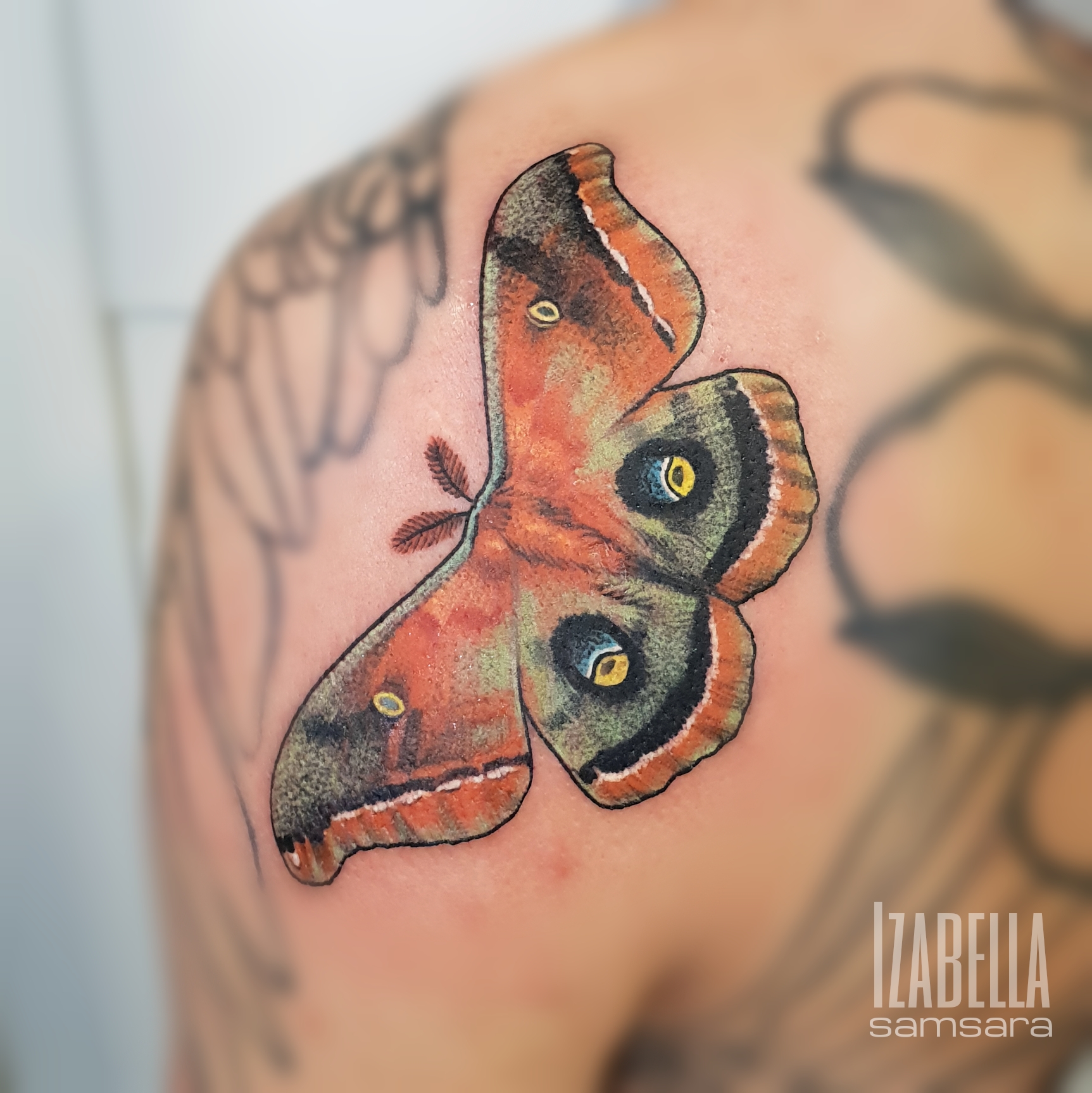 A colorful tattoo of a butterfly on the shoulder, featuring intricate details with shades of orange, black, and yellow. The butterfly has large, striking wings adorned with vivid patterns and eyespots, giving it a realistic and vibrant appearance.