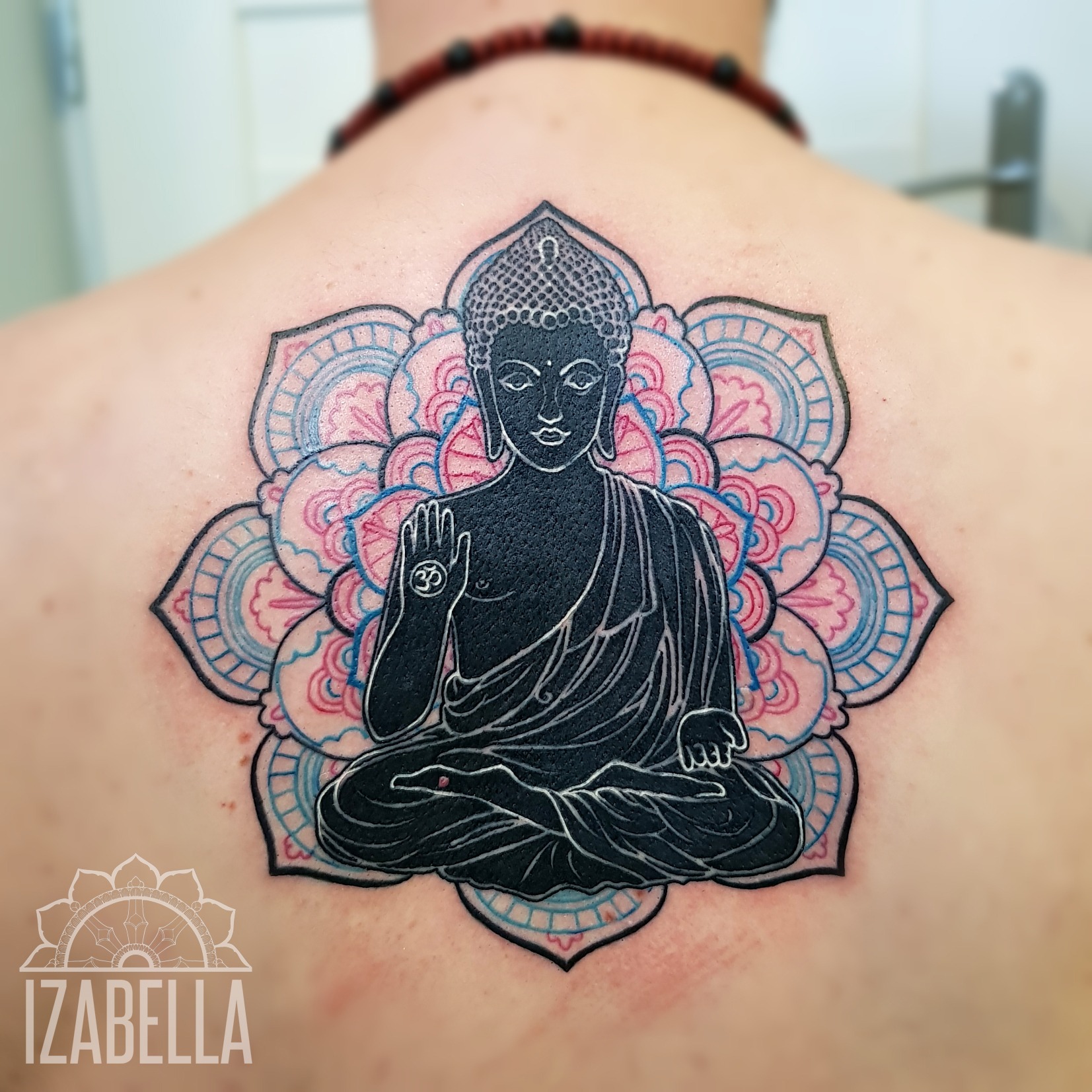 A detailed tattoo of a seated Buddha figure in a meditative pose, centered within a vibrant mandala design. The Buddha is depicted in dark ink, with intricate line work and a serene expression, while the surrounding mandala features soft pink and blue hues, enhancing the overall spiritual and artistic vibe of the tattoo. This artwork covers a significant area on the person's back.