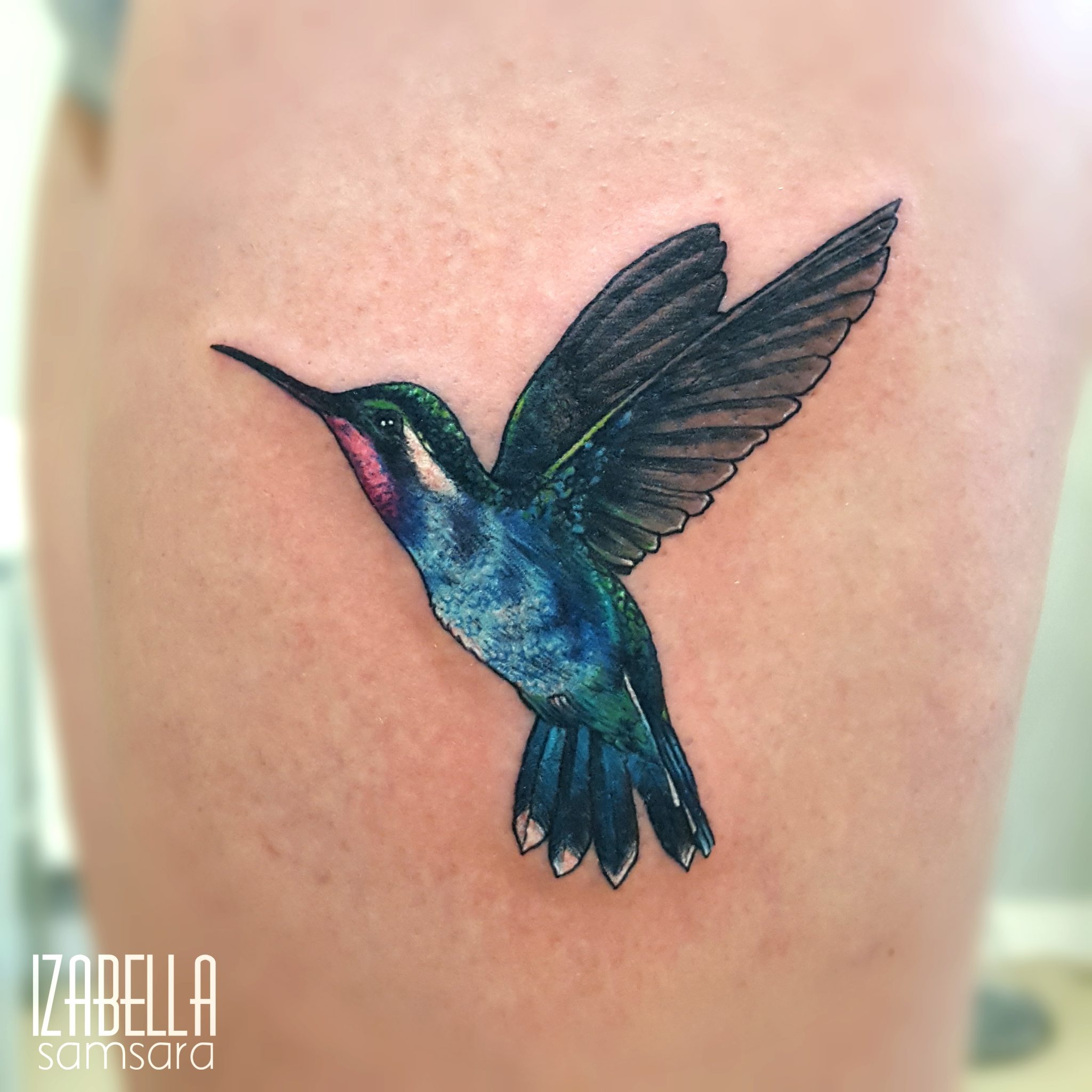 A vibrant tattoo of a hummingbird in flight, featuring detailed feathers in shades of blue, green, and purple. The tattoo showcases the bird's distinctive long beak and outstretched wings, set against a smooth skin background.