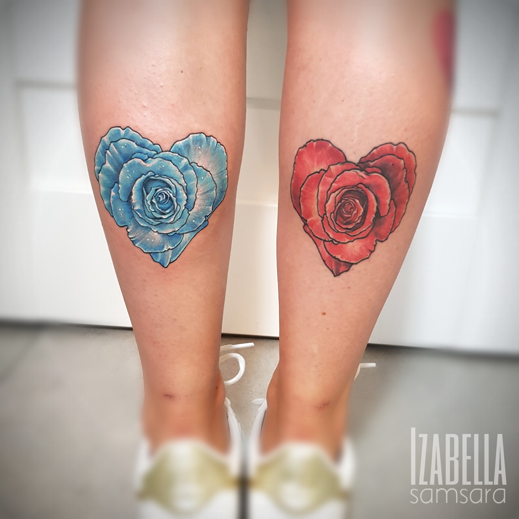 A vibrant tattoo featuring two heart-shaped roses on the calves, one in blue and the other in red. The blue rose is detailed with shades that create depth and dimension, while the red rose has a rich color with intricate shading, both designed to symbolize love and passion.