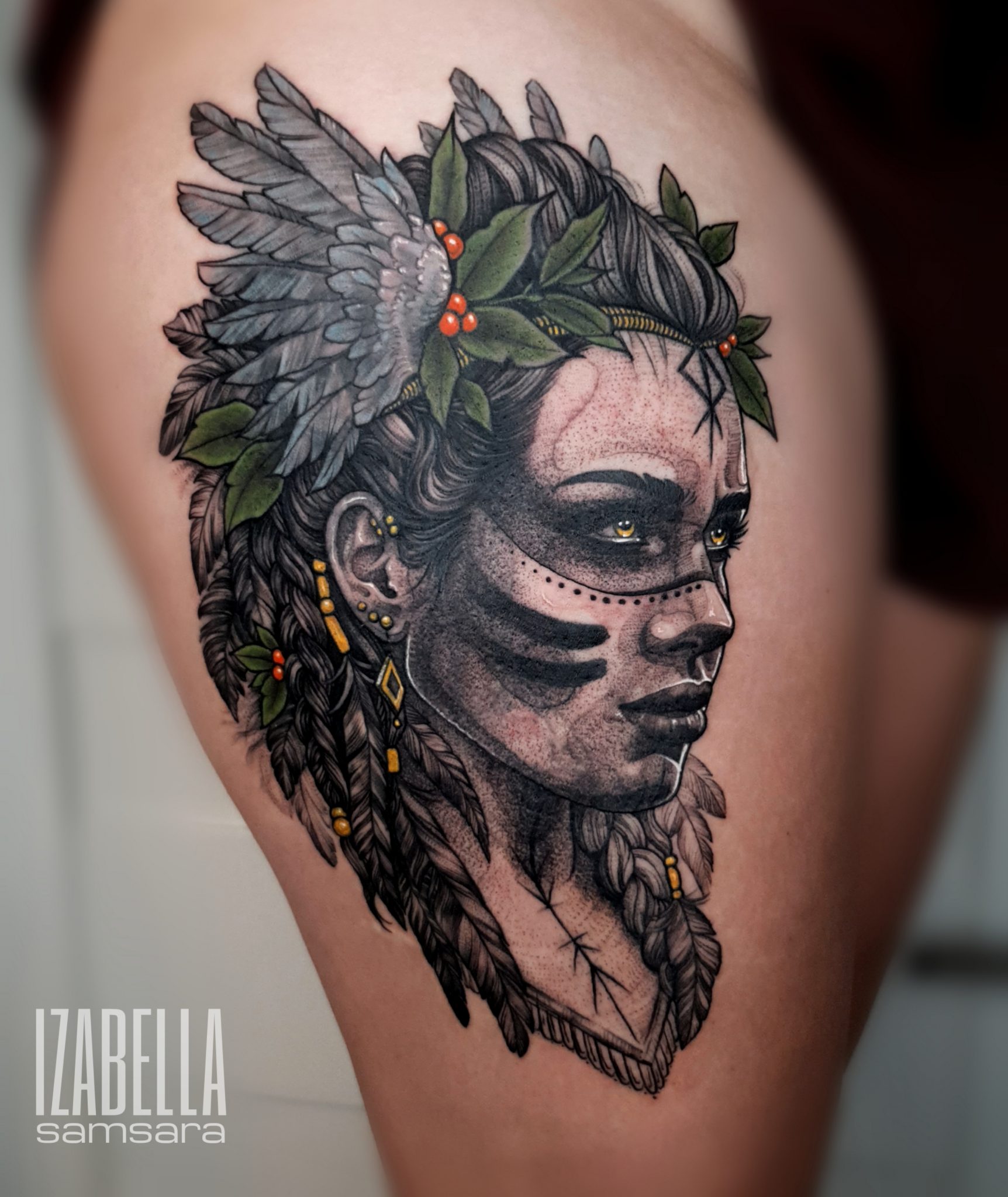 Black and gray tattoo featuring a detailed portrait of a woman with tribal face paint and decorative hair elements. The design includes feathers, leaves, and ornaments, showcasing intricate line work and shading.