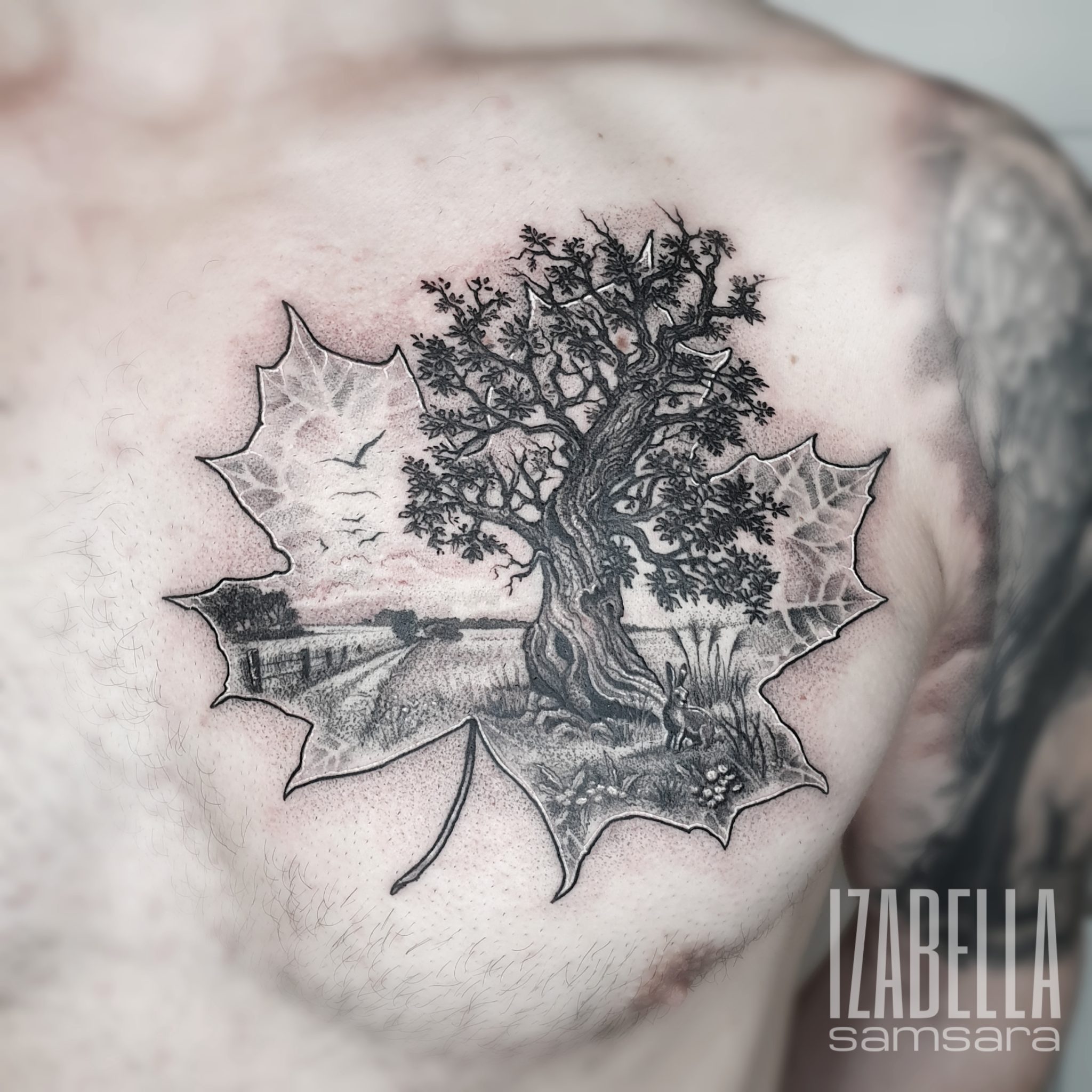 A detailed black and gray tattoo featuring a large oak tree with intricate branches and leaves, set within the outline of a maple leaf. In the background, there is a serene landscape with a field, a distant house, and a sky with birds flying. A small rabbit can be seen in the foreground among grass and flowers, creating a natural, peaceful scene.