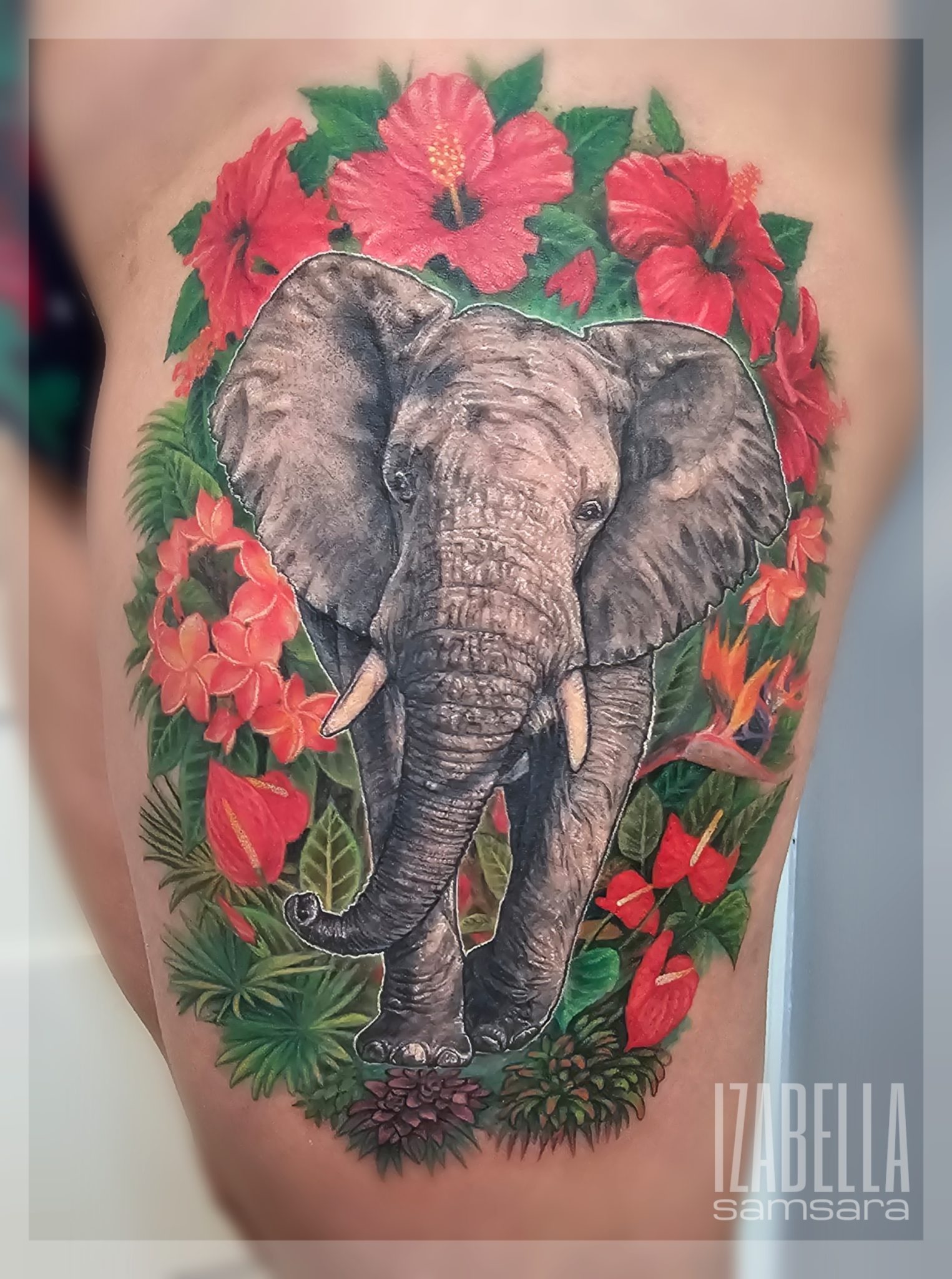 A vibrant tattoo featuring a realistic elephant surrounded by tropical flowers, including hibiscus and orchids, set against lush green foliage. The elephant has detailed textures and shading, enhancing its lifelike appearance. This colorful piece is ideal for nature lovers and those seeking animal-themed tattoos.