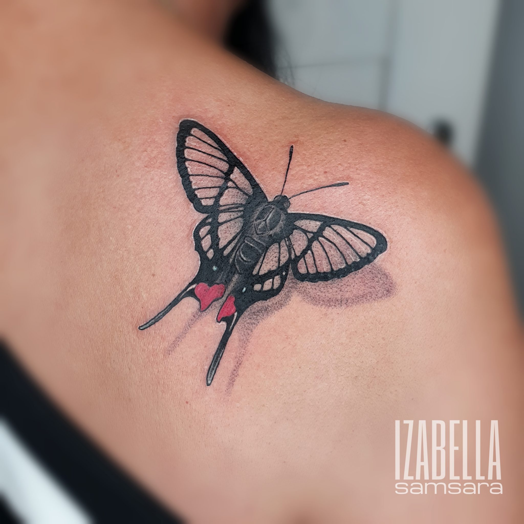 A beautifully detailed black and gray tattoo of a butterfly with intricate wing patterns and subtle shading. The butterfly features small vibrant red hearts near its body, adding a splash of color to the overall monochromatic design. This tattoo is placed on the shoulder, showcasing its elegant form and fine line work.