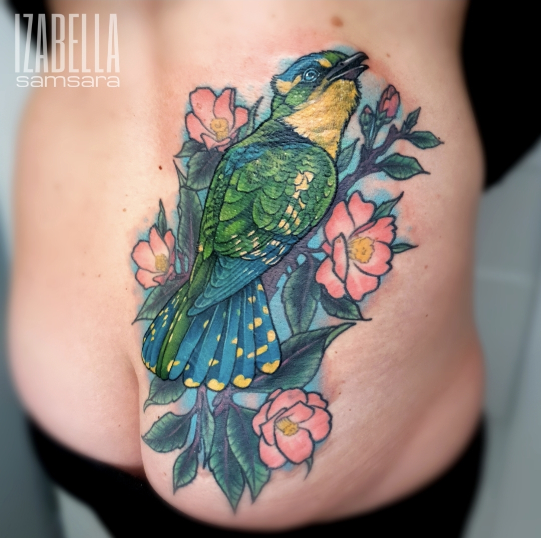 A vibrant tattoo of a bird perched among pink flowers and green leaves, featuring detailed shading and bright colors. The bird has a green and blue plumage with a yellow underside and is singing, surrounded by delicate floral elements, ideal for nature and bird enthusiasts.