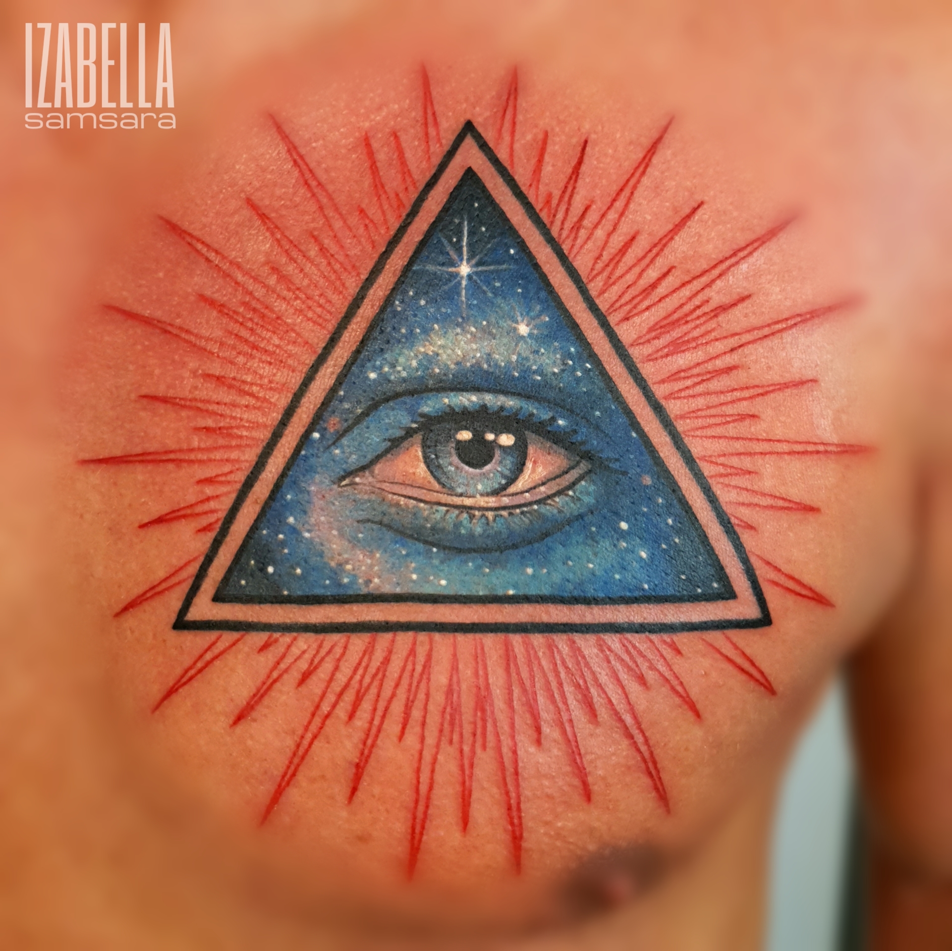 A vibrant tattoo featuring a detailed eye within a blue triangle, surrounded by radiant red beams. The eye is expressive and depicts a cosmic scene, adding depth to the design. The tattoo symbolizes perception and enlightenment, with a beautifully blended color palette.