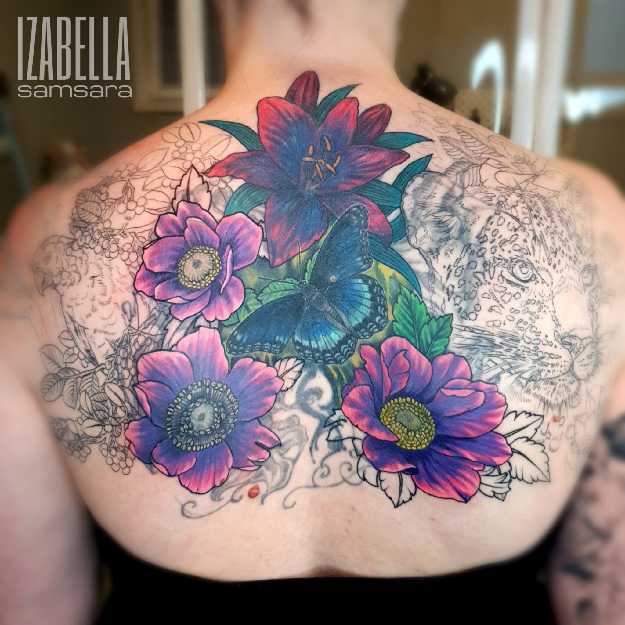 Vibrant back tattoo featuring colorful flowers including purple lilies and anemones, with a detailed blue butterfly at the center. The design also includes intricate line work of a leopard and various floral elements, creating a striking blend of nature and artistry.