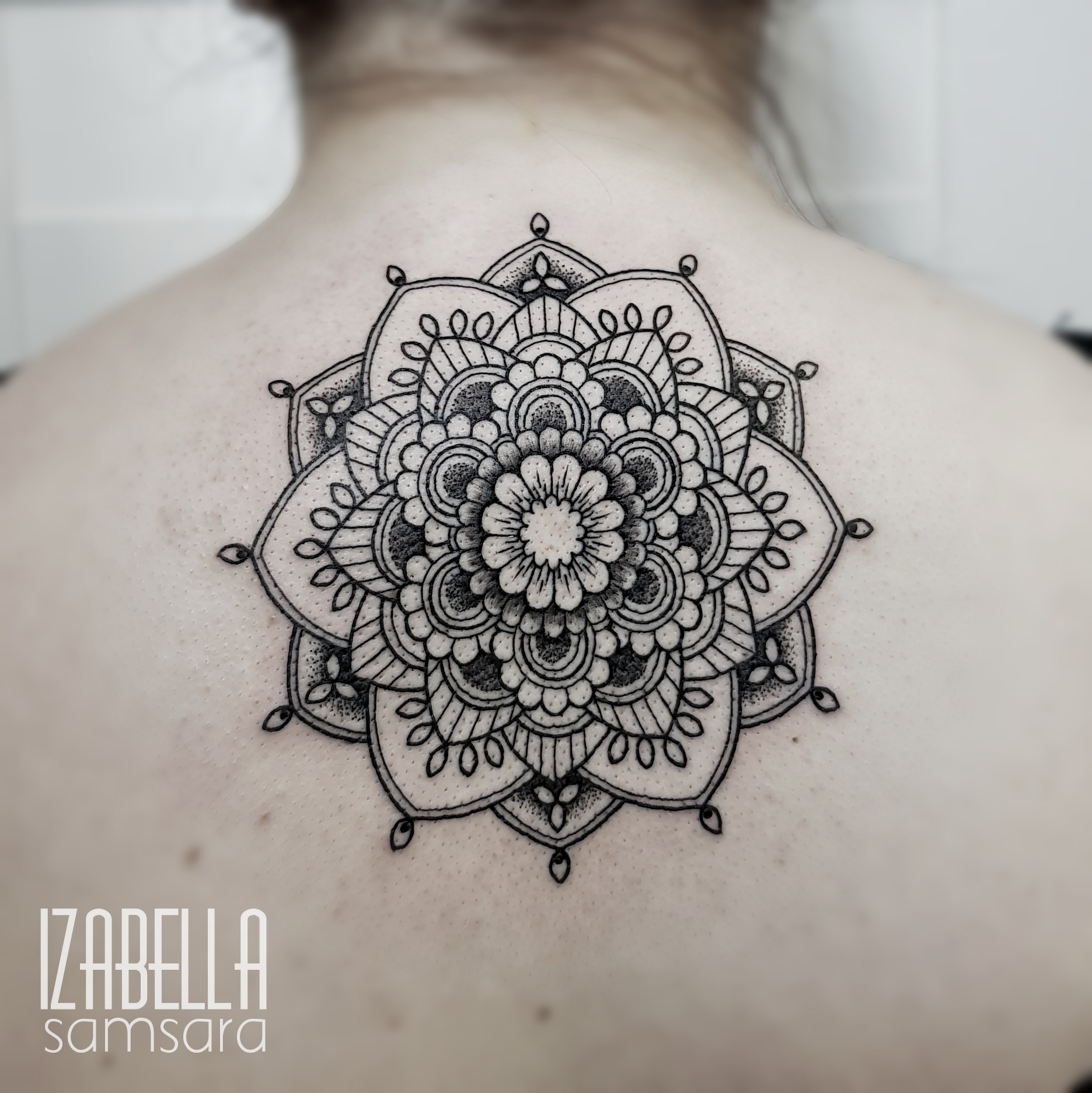 A detailed black and white tattoo featuring a mandala design on the upper back. The intricate artwork showcases multiple layers of floral patterns, geometric shapes, and symmetrical elements, creating a visually striking appearance that emphasizes the artistry of linework and dot shading.