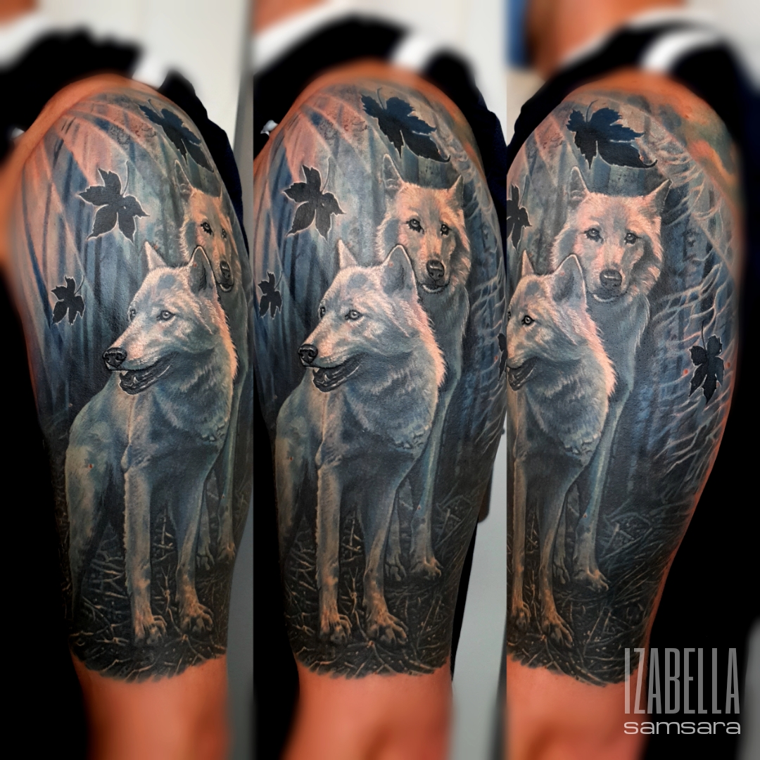 A detailed black and grey tattoo featuring two wolves standing side by side in a natural setting. The wolves have realistic fur texture and expressions, surrounded by foliage and gentle shadows. The artwork captures a serene atmosphere, emphasizing the wolves' eyes and features, creating a lifelike appearance.