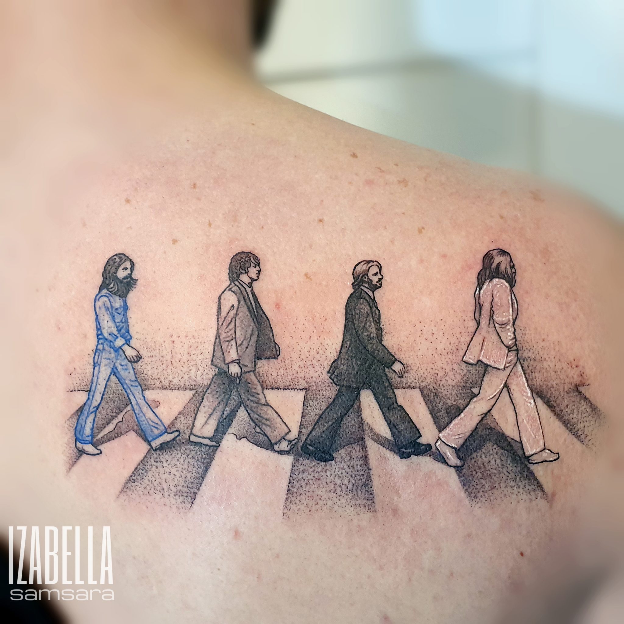 A colorful tattoo of four iconic figures, depicted in a walking motion, inspired by the famous Abbey Road album cover. The first figure wears a blue suit, followed by three in varying shades of black and white suits, walking across a pedestrian crossing. The design incorporates fine details, shading, and a gradient effect, creating a striking visual representation of the band.