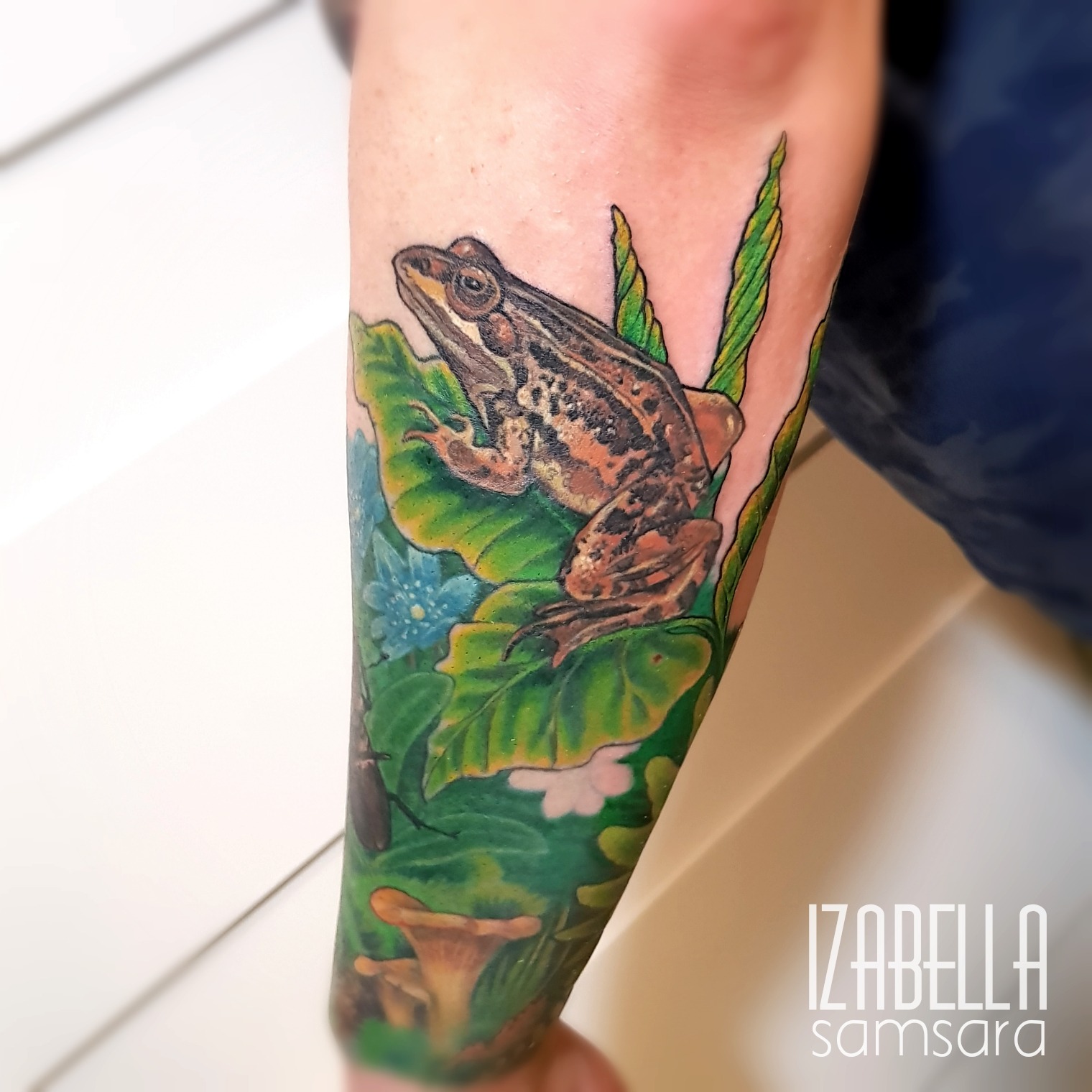 A detailed tattoo featuring a realistic frog perched on vibrant green leaves, surrounded by colorful flowers and foliage. The design showcases intricate shading and vivid colors, capturing the natural beauty of the frog in a dynamic pose.