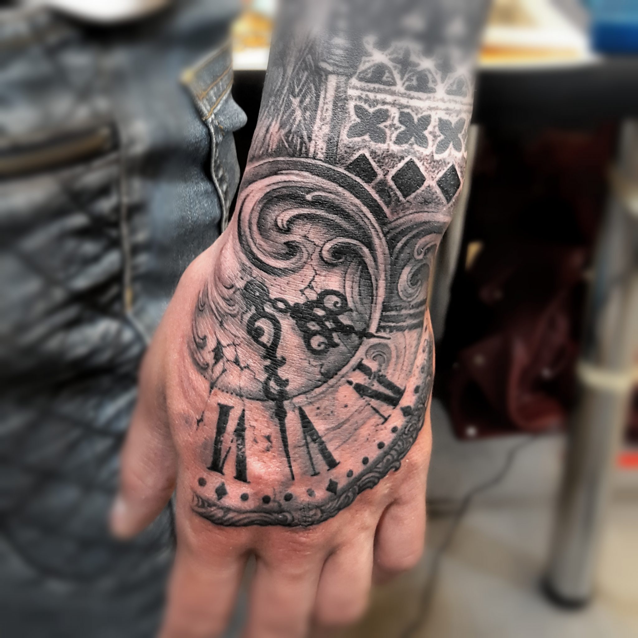 A detailed black and gray tattoo covering the hand and wrist, featuring an ornate clock design with Roman numerals, intricate swirls, and decorative elements. The shading creates a realistic effect, showcasing the craftsmanship of the tattoo artist.