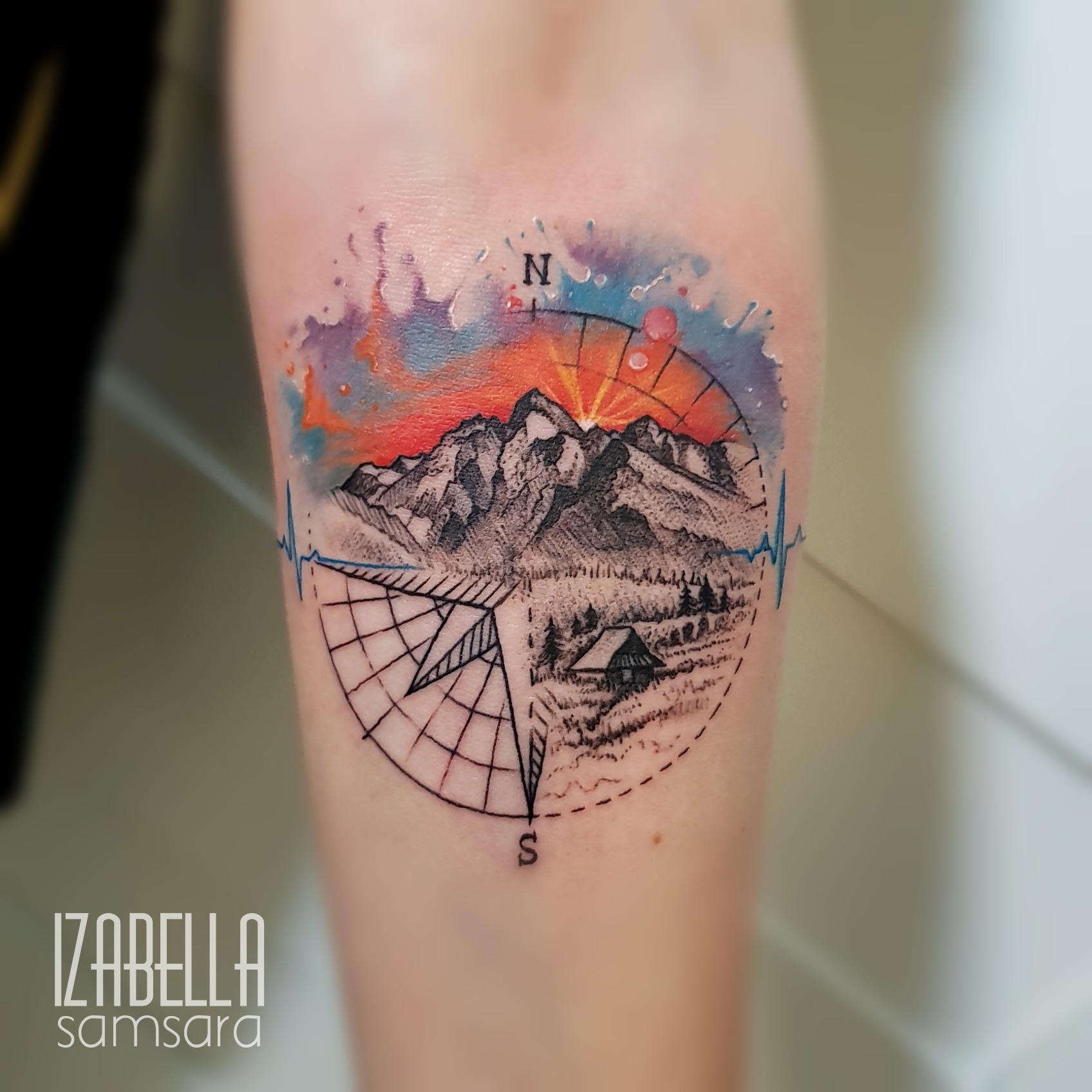 A colorful tattoo featuring a mountain landscape with a vibrant sunset and abstract watercolor splashes in orange and purple. It includes a detailed compass design with a North indicator and a grid pattern, alongside elements like a small cabin and tree line. The artwork combines fine line work and vivid colors, creating a dynamic and adventurous theme.