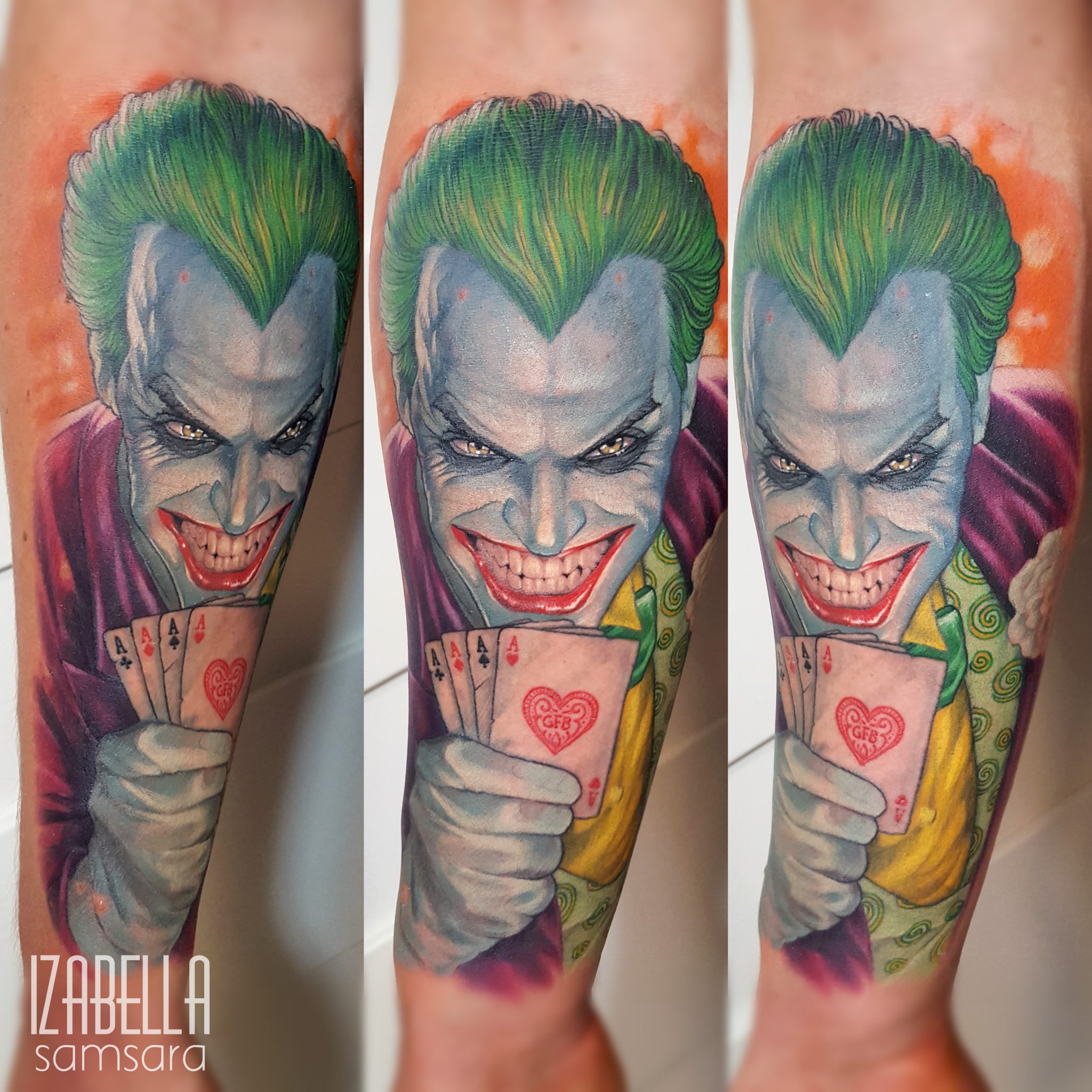 A vibrant, colorful tattoo depicting the Joker character from comic books. The Joker is shown with green hair, a wide grin, and black eyes, holding playing cards in a menacing pose. His outfit features purple and green colors, with intricate patterns and details. The background is a blend of orange hues, enhancing the dramatic effect of the tattoo.
