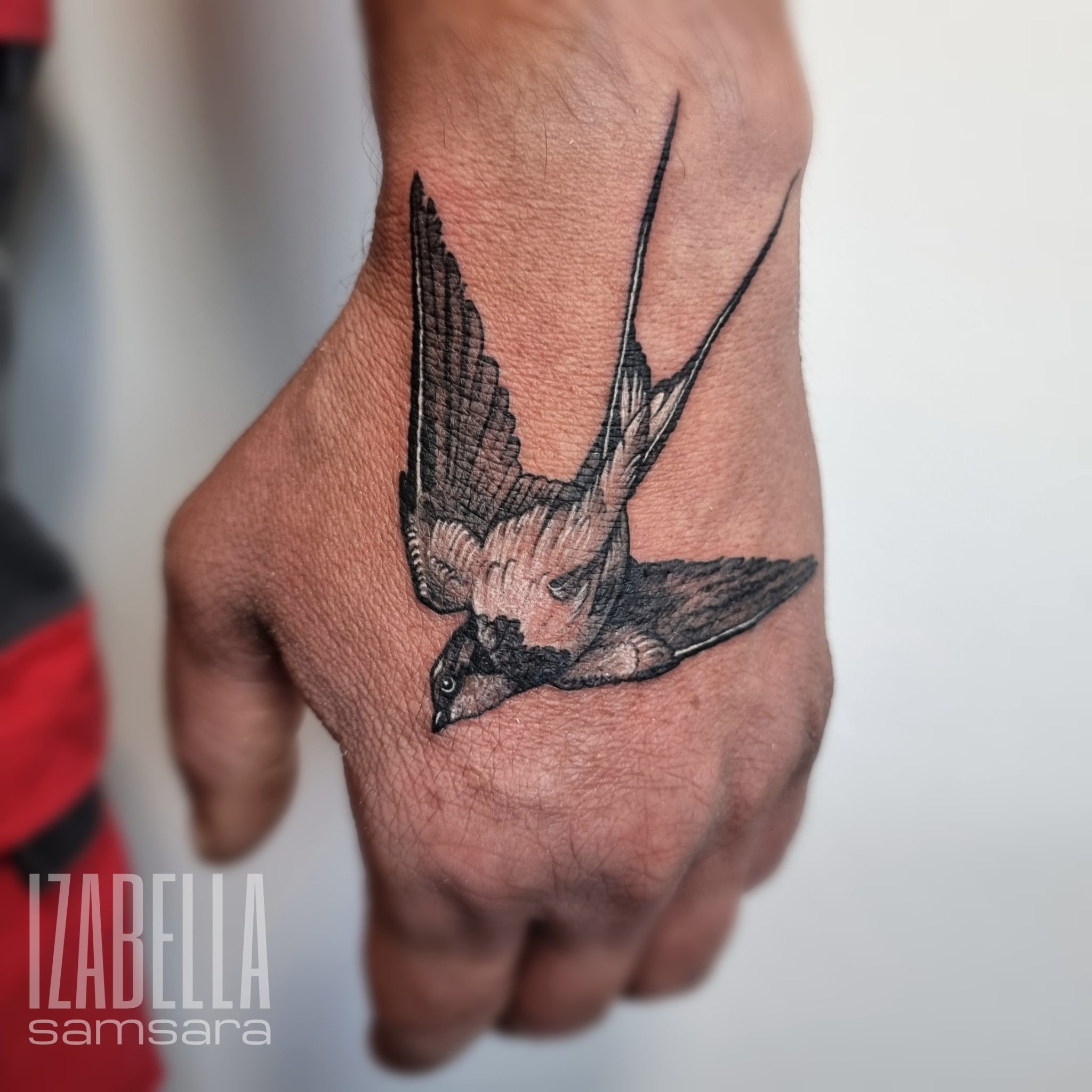 A detailed black and gray tattoo of a flying bird, specifically a swallow, depicted on a hand. The tattoo showcases the bird in an upward position with its wings spread wide, featuring intricate shading and lifelike detailing. The hand's skin tone contrasts with the dark ink, enhancing the tattoo's visibility and artistic depth.