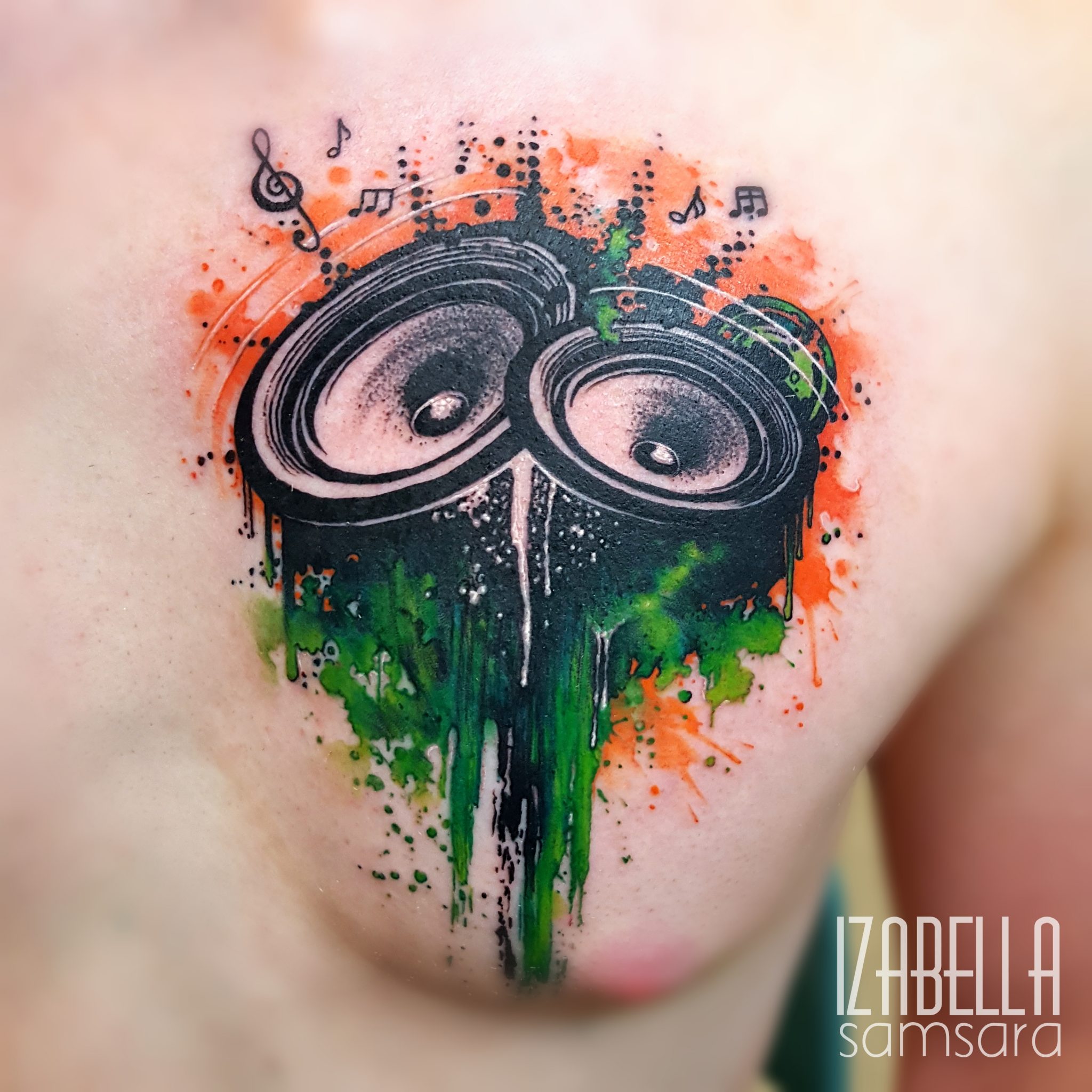 A vibrant tattoo featuring two speaker cones, surrounded by colorful splashes of orange and green ink. The design includes musical notes and a dripping paint effect, creating an energetic and artistic expression of music and sound.