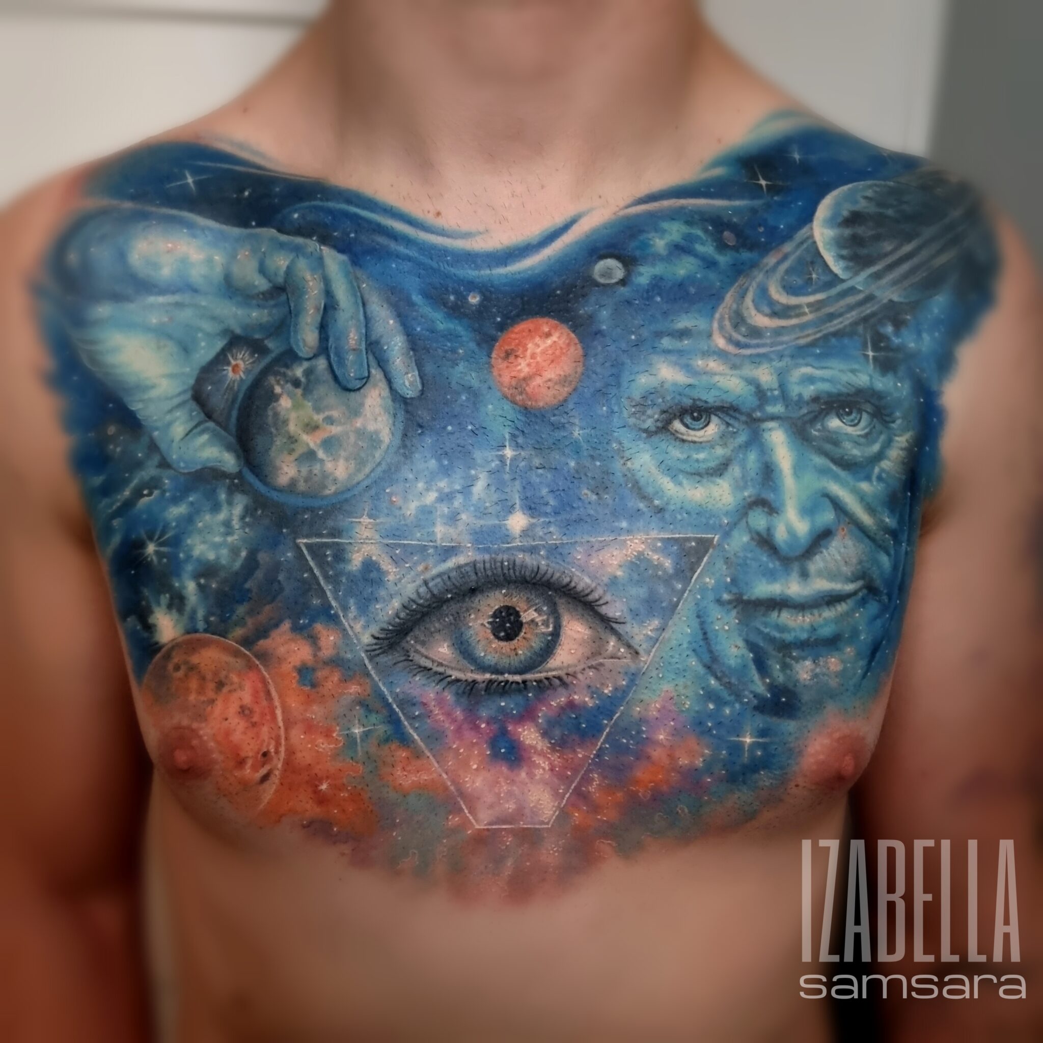 A vibrant tattoo covering the chest, featuring a cosmic scene with planets and galaxies. Central elements include a detailed human eye inside a triangle, a mysterious blue face, and a hand holding a globe, all intricately shaded in blues, greens, and reds, depicting the vastness of space and a sense of wonder.
