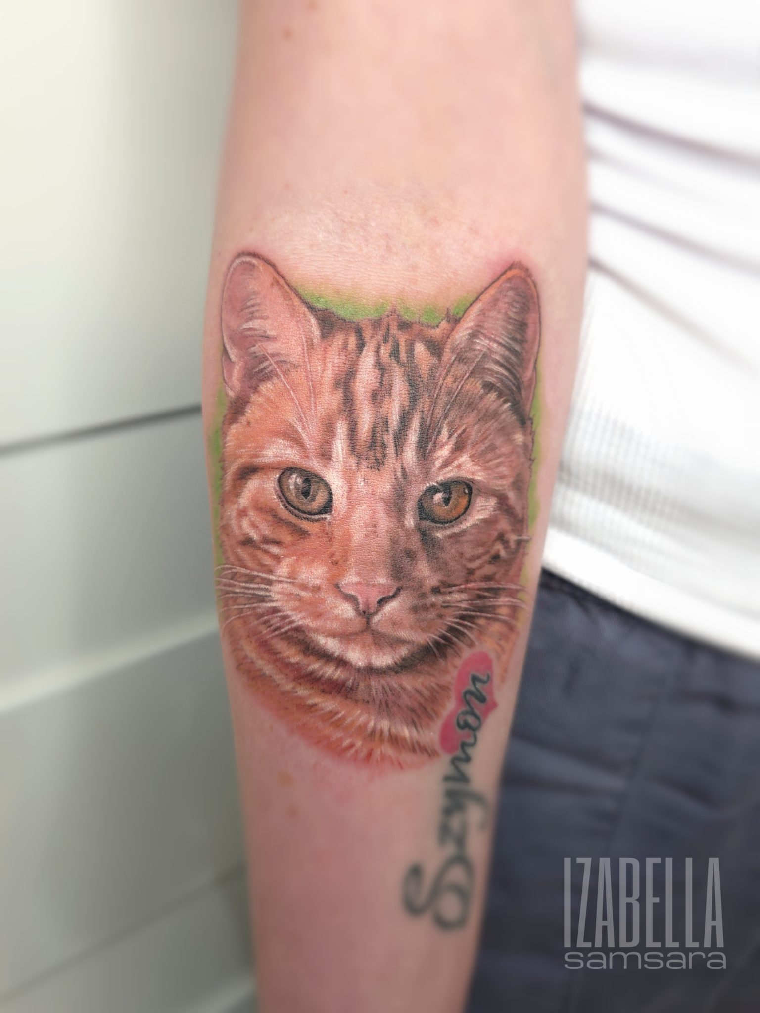 Realistic tattoo of a tabby cat's face, showcasing detailed fur patterns and expressive eyes, with a green background. The tattoo also features the name 'Szymon' in a stylish script below the cat.