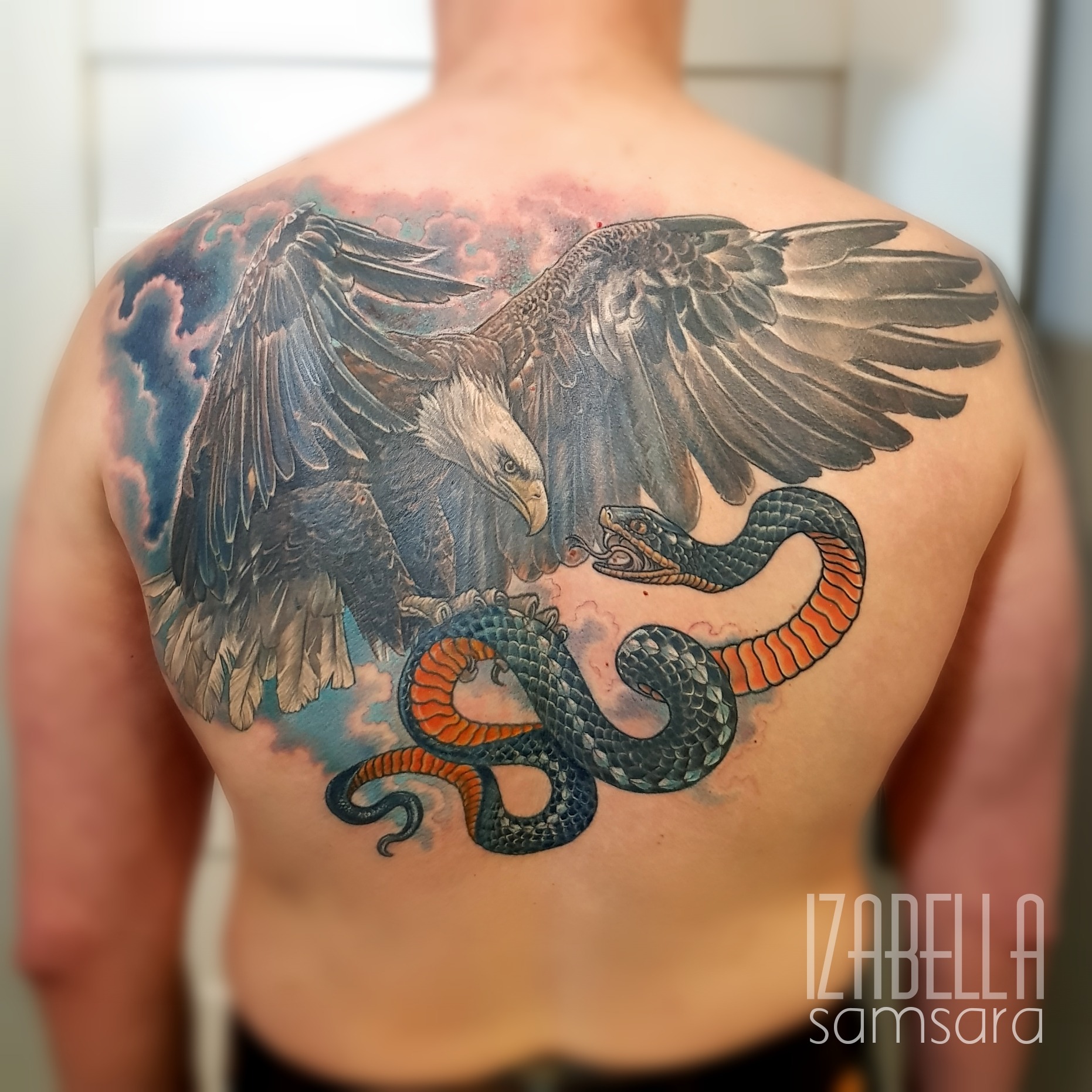 A stunning full back tattoo featuring a fierce eagle with outstretched wings soaring above a coiling snake. The eagle's detailed feathers are accentuated with shades of gray and black, while the snake showcases vibrant hues of green and orange. The background is adorned with swirling clouds, creating a dramatic sky effect. This colorful tattoo beautifully captures the dynamic relationship between the eagle and the snake.