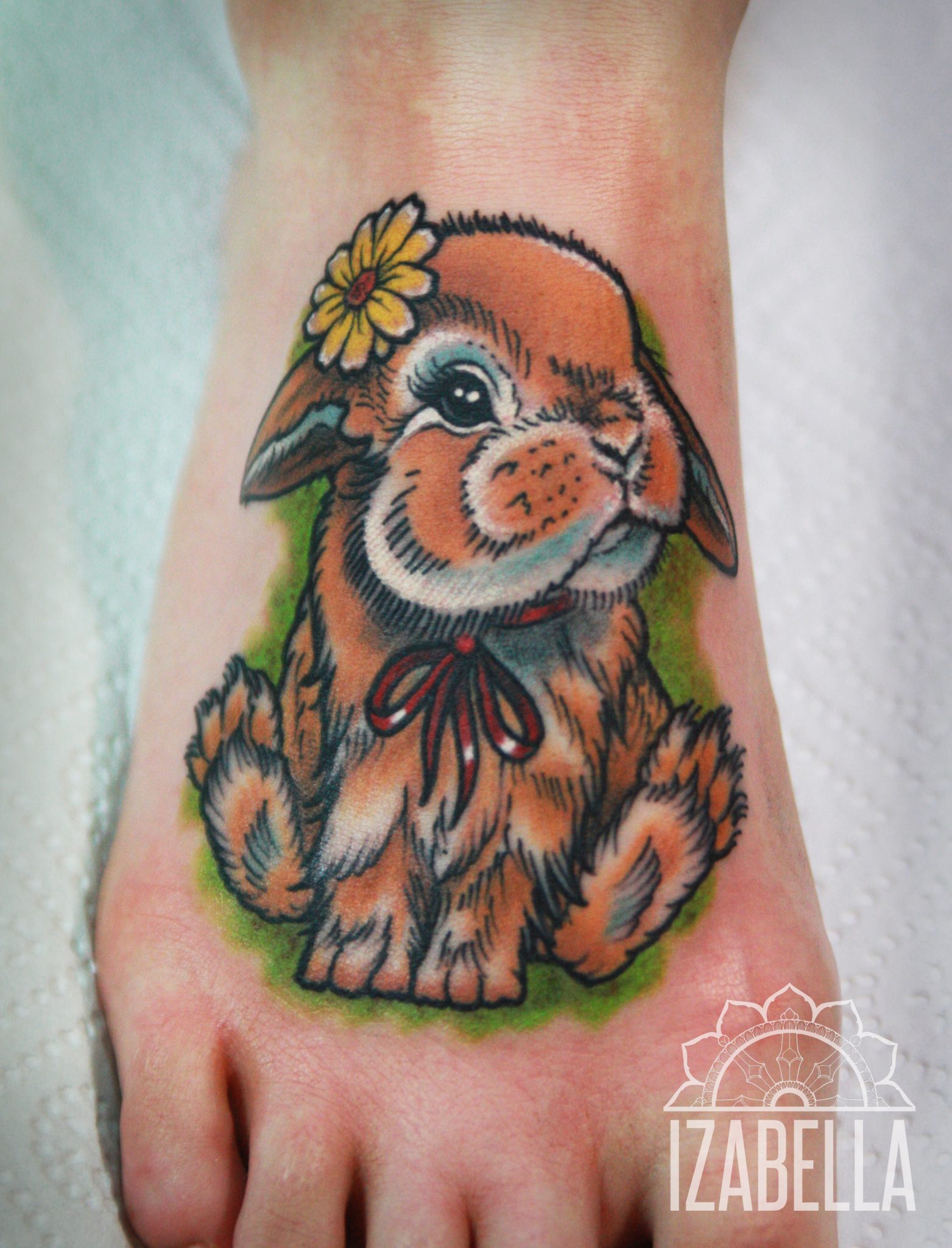 A colorful tattoo of a cute, fluffy rabbit sitting with a cheerful expression. The rabbit has a flower tucked behind its ear and is adorned with a red ribbon around its neck. The background features soft green shading that adds depth to the design, making the rabbit stand out vividly.