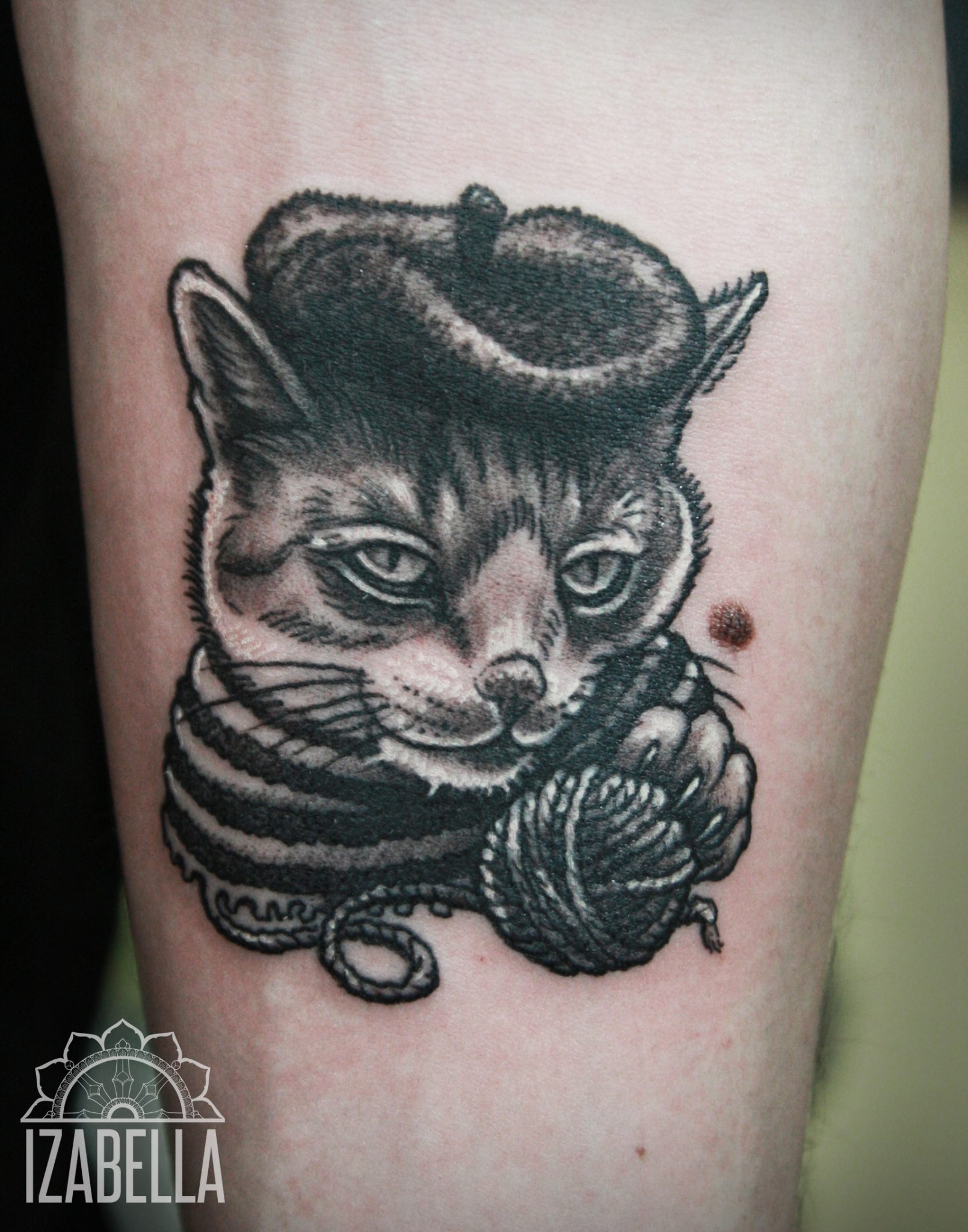 Black and white tattoo of a cat wearing a beret and a striped scarf, playfully holding a ball of yarn. The cat has detailed features, with expressive eyes and whiskers, showcasing realistic shading and intricate line work.