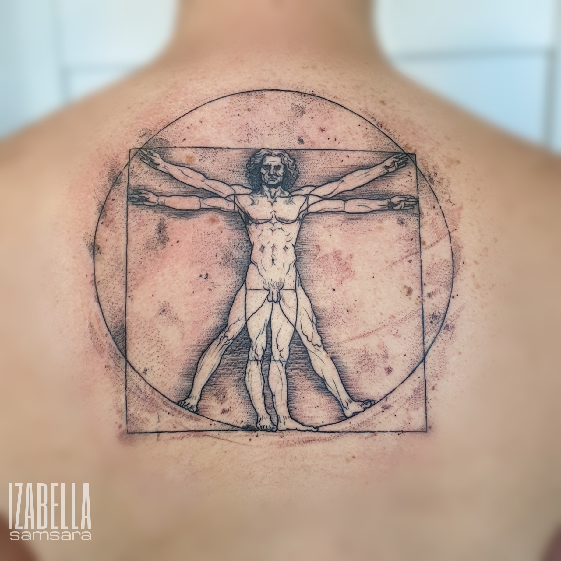 A detailed black and white tattoo depicting the Vitruvian Man by Leonardo da Vinci, featured on the upper back. The design includes both a circle and a square around the figure, showcasing the ideal human proportions with outstretched arms and legs, blended meticulously into the skin.