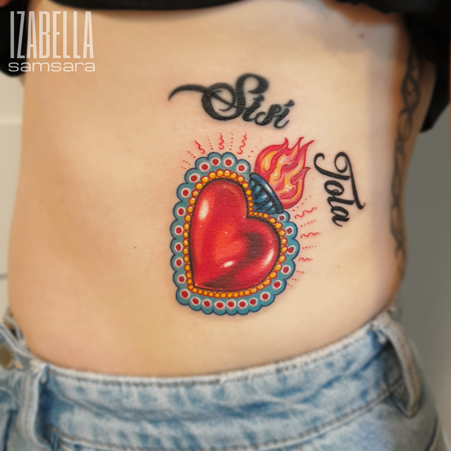 A vibrant tattoo featuring a large red heart with a flame emerging from the top, surrounded by a decorative blue and gold border. Above the heart, the word 'Siost' is elegantly scripted, and below it, the name 'Tota' is also displayed in a stylish font. This colorful design is positioned on the ribcage, combining elements of traditional and modern tattoo artistry.