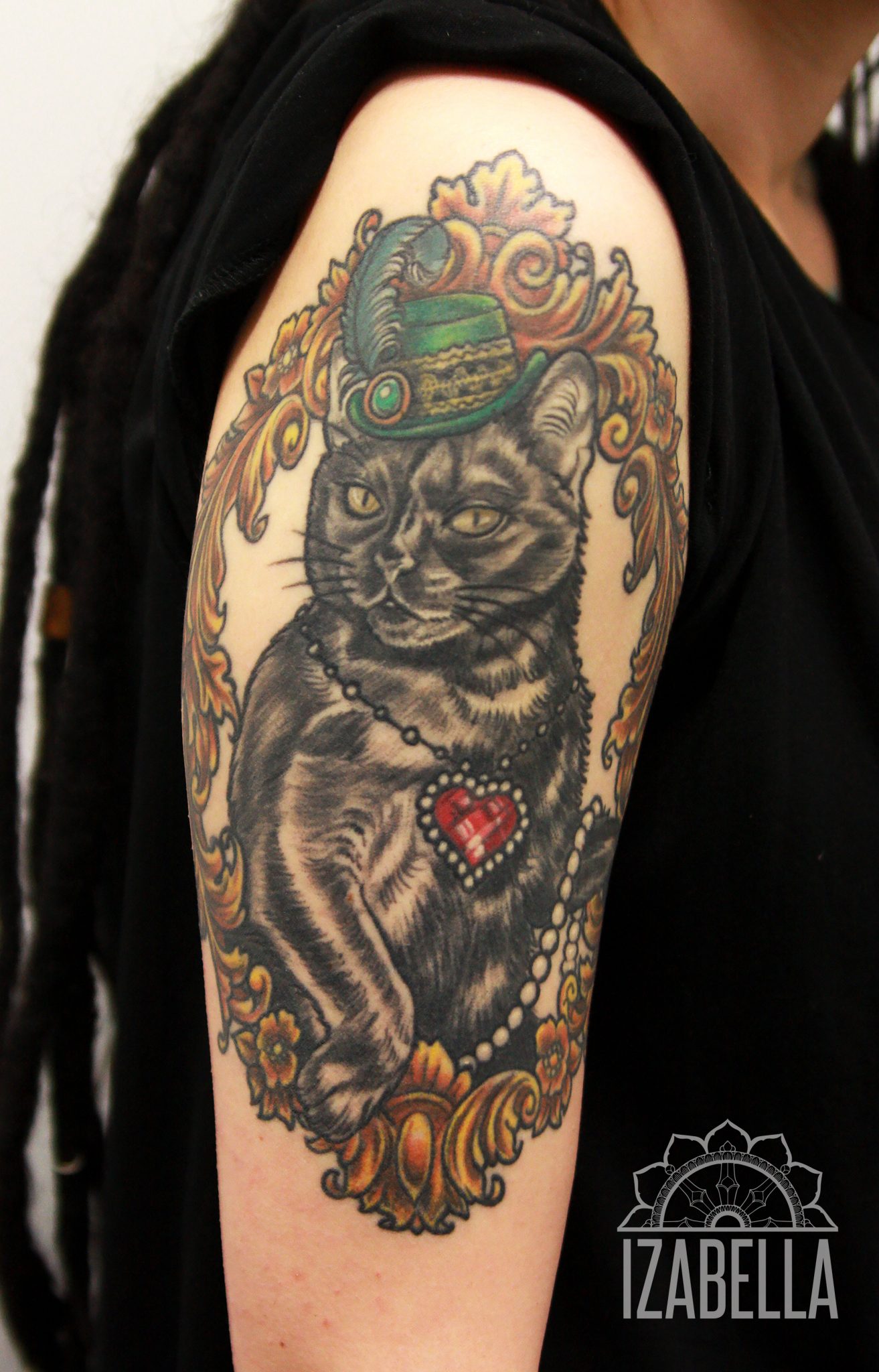 Tattoo of a stylish black cat wearing a green hat with a feather, adorned with a pearl necklace and a heart-shaped locket. The cat is framed by ornate decorative elements, featuring flowers and intricate patterns, creating an elegant and whimsical design.