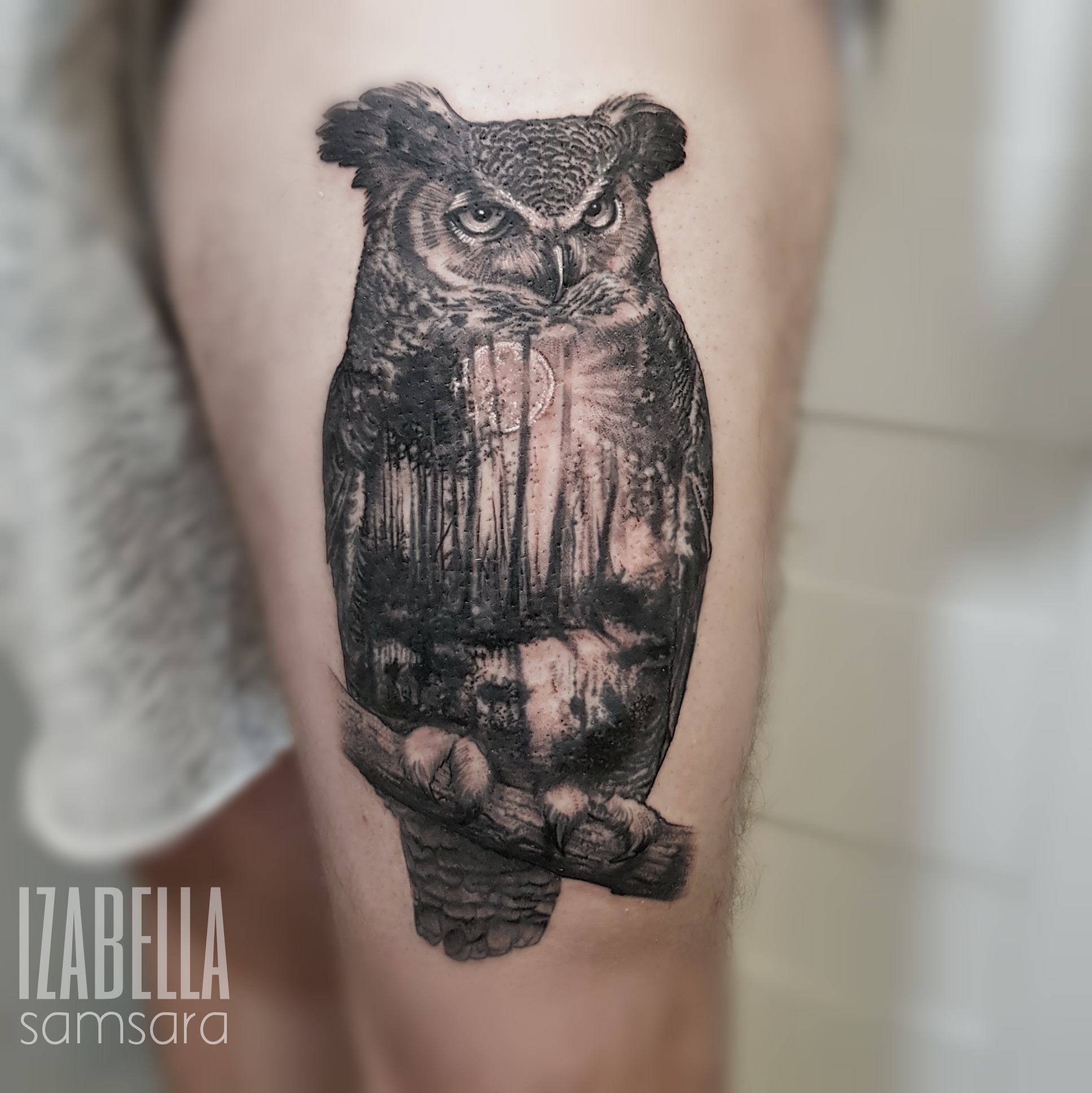 A detailed black and grey tattoo of an owl perched on a branch, featuring intricate feather patterns and a piercing gaze. The background incorporates elements of a forest, delivering a mystical ambiance. This realistic tattoo showcases artistic shading and fine lines, highlighting the owl's majestic presence.