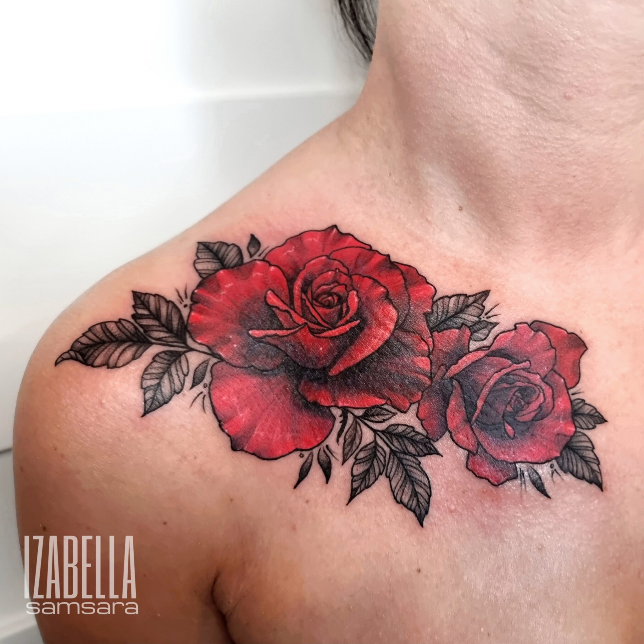 A vibrant tattoo of two red roses with detailed petals and green leaves, located on the upper shoulder and neckline. The flowers are intricately designed, showcasing the depth of color and shading, creating a lifelike appearance.