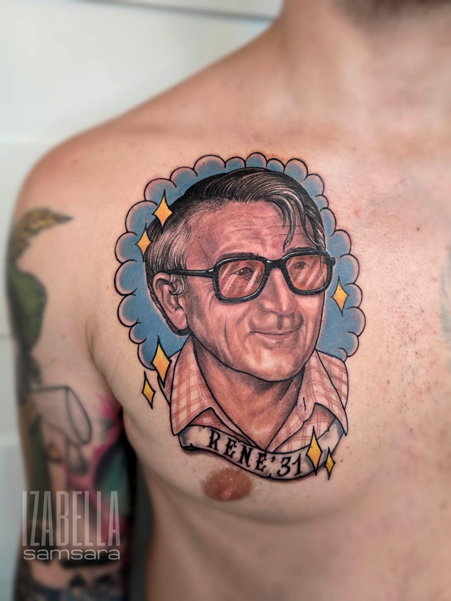 A colorful tattoo depicting a portrait of a man with glasses, styled in a traditional art form. The man is shown smiling, wearing a plaid shirt. Surrounding the portrait is a decorative blue cloud with sparkling stars. Below the portrait, a banner reads 'RENE' with the number '31.' This chest tattoo exudes a nostalgic and vintage vibe, perfect for honoring loved ones.