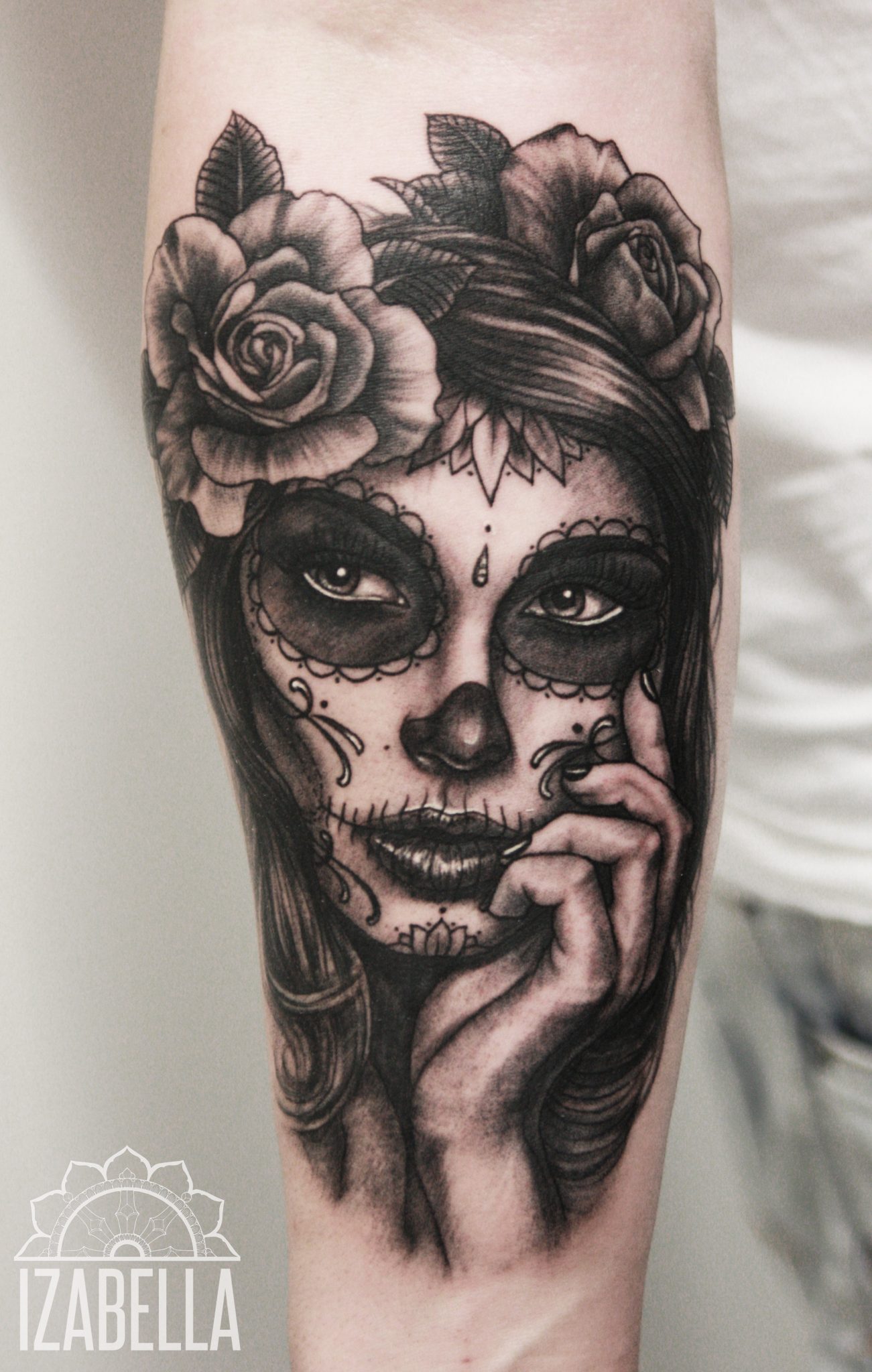 A detailed black and gray tattoo of a sugar skull girl with intricate designs on her face, surrounded by blooming roses. The girl has long flowing hair and is holding her chin with one hand, showcasing a captivating expression with bold eye makeup and decorative elements like dots and swirls on her face.