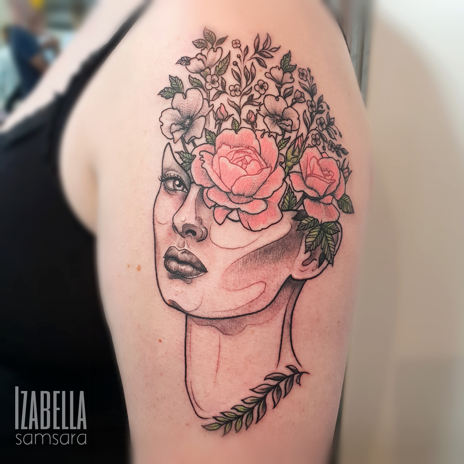 A striking tattoo featuring a stylized, abstract face adorned with vibrant roses and delicate floral elements. The design includes intricate line work, blending black and shades of pink, showcasing a balance of nature and artistry. The tattoo is placed on the upper arm, emphasizing the graceful contours of the face and the lush floral arrangement.