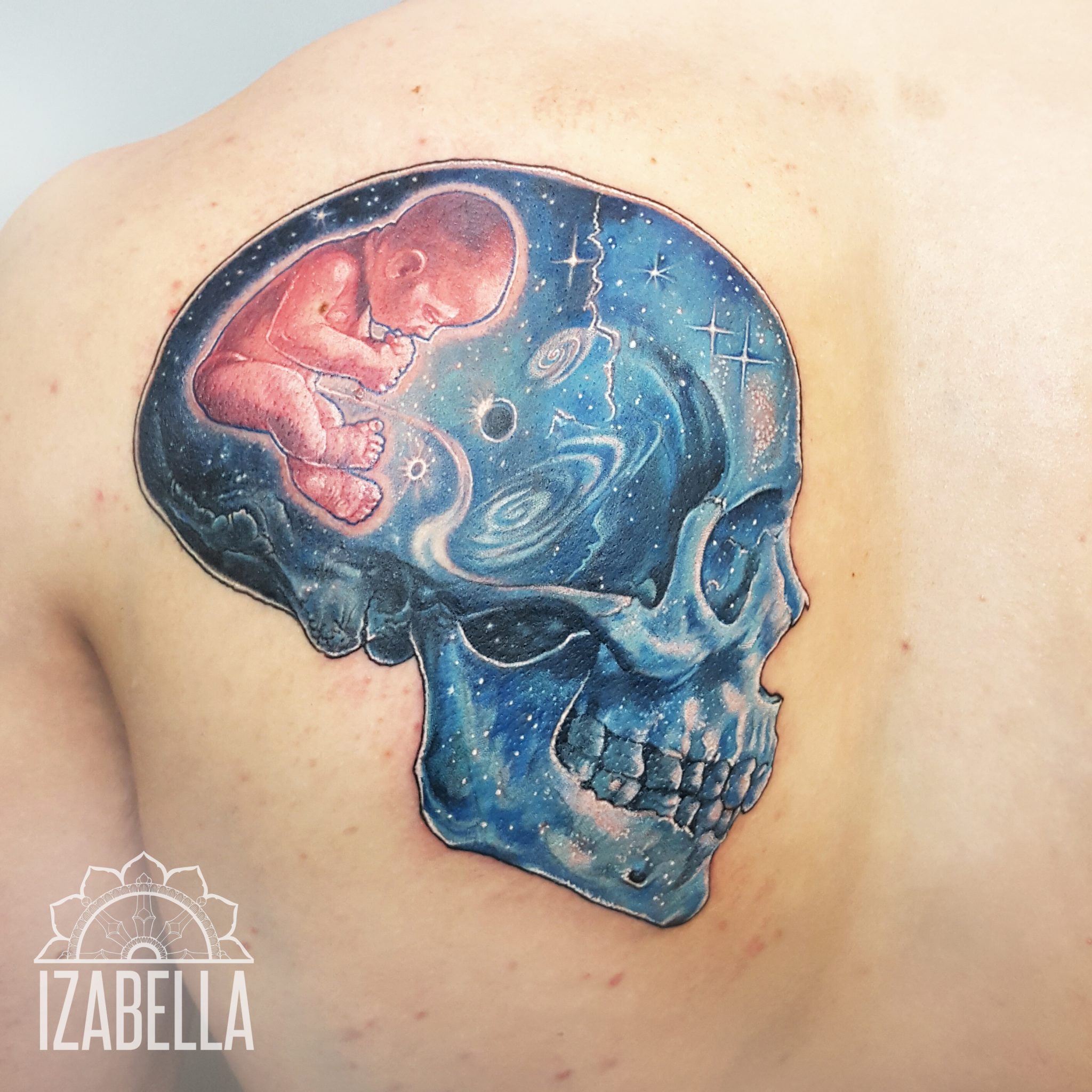 A colorful tattoo depicting a human skull with a galaxy and space theme. Inside the skull, there is a representation of a fetal baby curled up, surrounded by stars and cosmic swirls, blending elements of life and death. The skull is primarily blue with intricate details, while the fetus is shown in shades of pink, creating a striking contrast.
