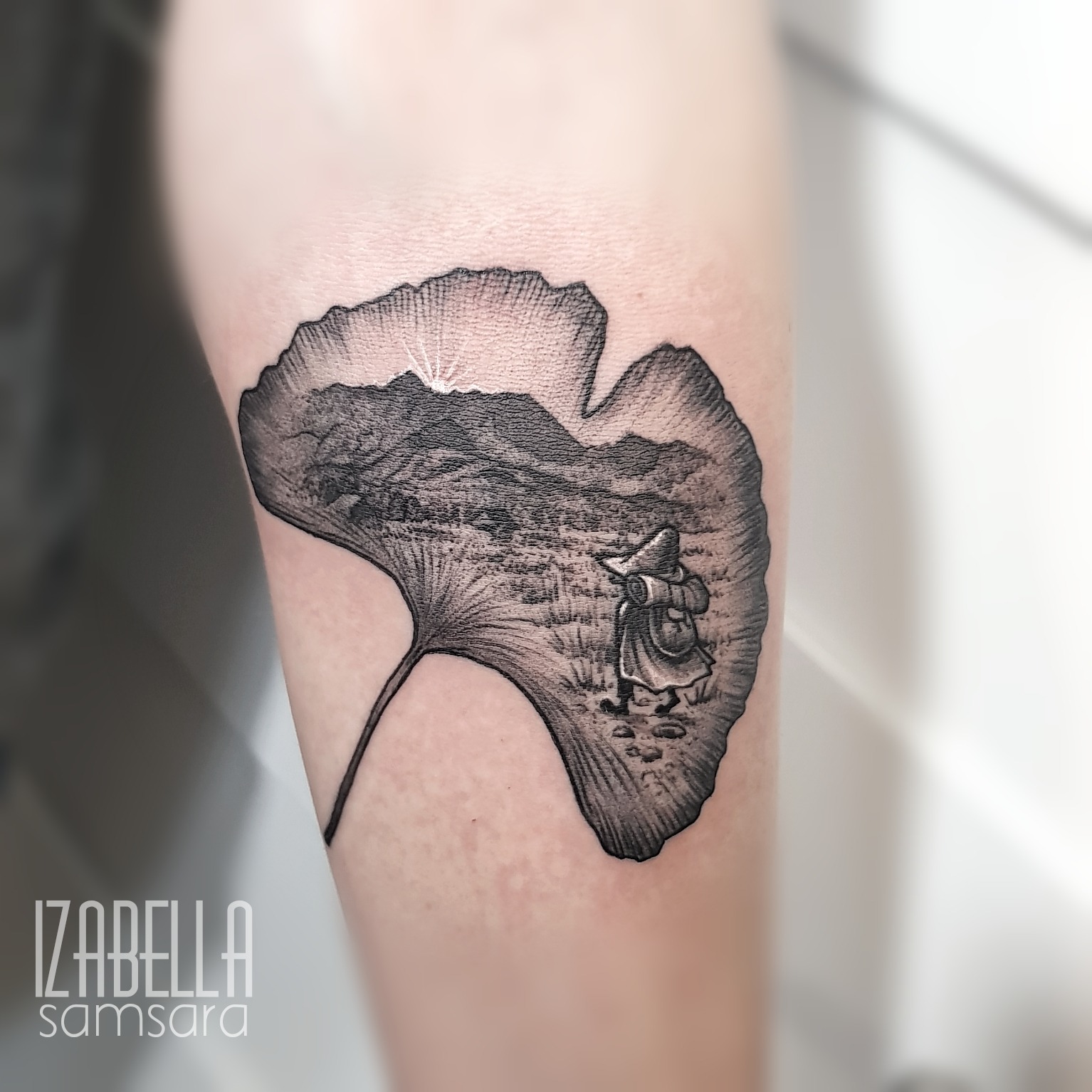 A black and grey tattoo depicting a ginkgo leaf shape that contains a scenic landscape. Inside the leaf, there is a detailed illustration of mountains and a small figure wearing a hat and carrying a bag, walking through the landscape. The design emphasizes depth and shading, creating a surreal effect, with sunlight breaking over the mountains in the background.