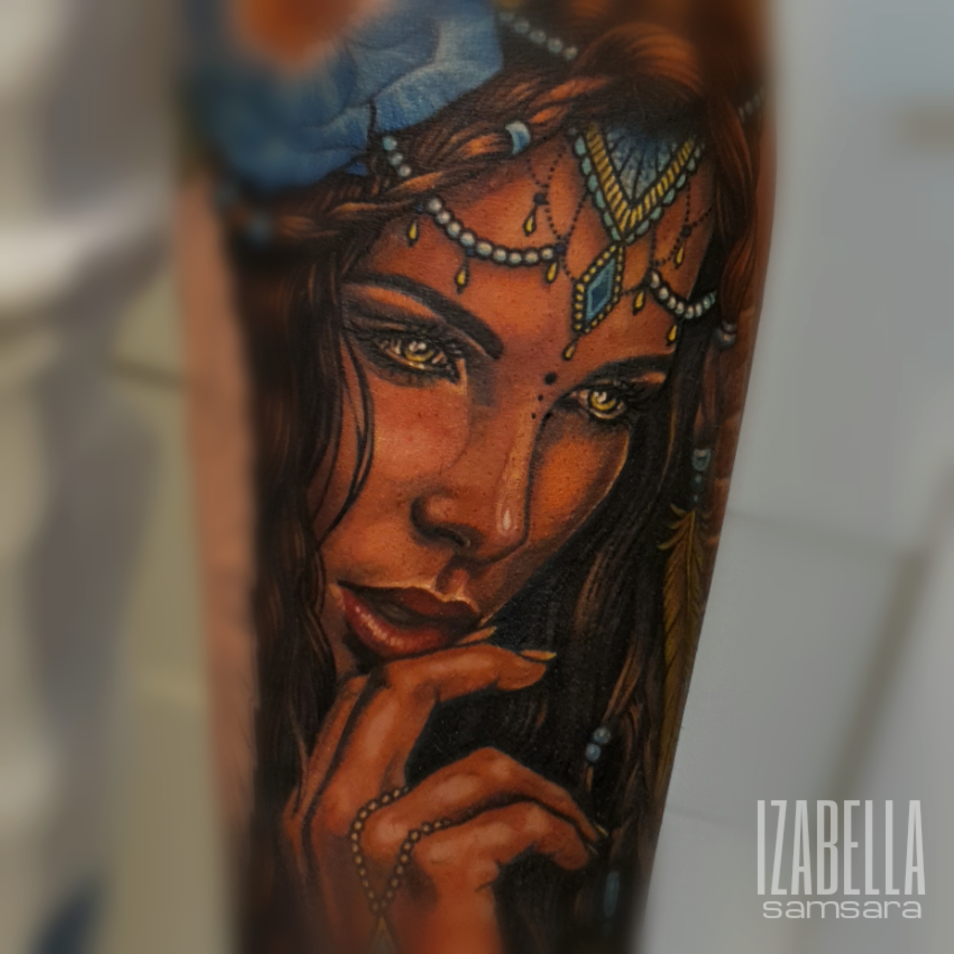 A vibrant tattoo depicting a woman's face with long, flowing hair, adorned with a blue flower and intricate jewelry. Her piercing golden eyes and detailed features are emphasized, while she gently touches her finger to her lips, creating a captivating and enchanting atmosphere.