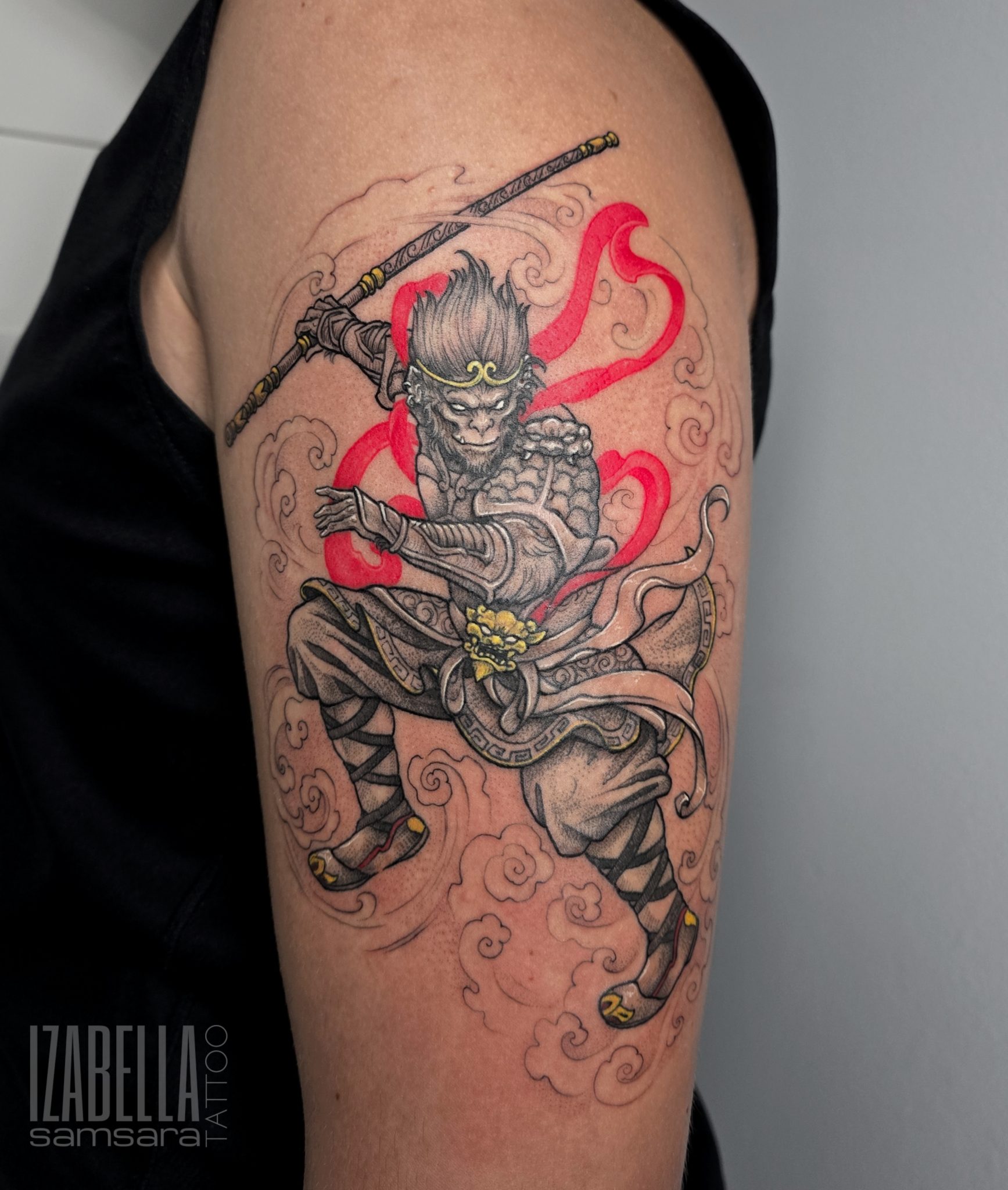 A vibrant tattoo depicting a warrior with monkey-like features, holding a staff and dressed in traditional armor with intricate details. The design includes swirling clouds and flowing red ribbons, emphasizing movement and action, showcasing a blend of artistry and cultural elements.